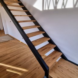 NORIX GM SPÓŁKA Z OGRANICZONĄ ODPOWIEDZIALNOŚCIĄ - Nowoczesne schody z drewnianymi stopniami i czarną metalową konstrukcją, widoczne na tle białej ściany z cieniem, uzupełnione drewnianą podłogą i belką stropową.