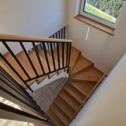 Spiralne stopnie z jasnego drewna z czarnymi metalowymi balustradami, widok z góry, z fragmentem buta w kadrze.