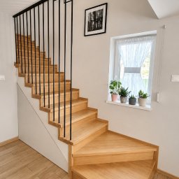 NORIX GM SPÓŁKA Z OGRANICZONĄ ODPOWIEDZIALNOŚCIĄ - Dębowe schody z czarną, strunową balustradą w nowoczesnym wnętrzu. Widoczne rośliny w oknie i drewnopodobna podłoga. Ujęcie z dołu, podkreślające solidność konstrukcji.