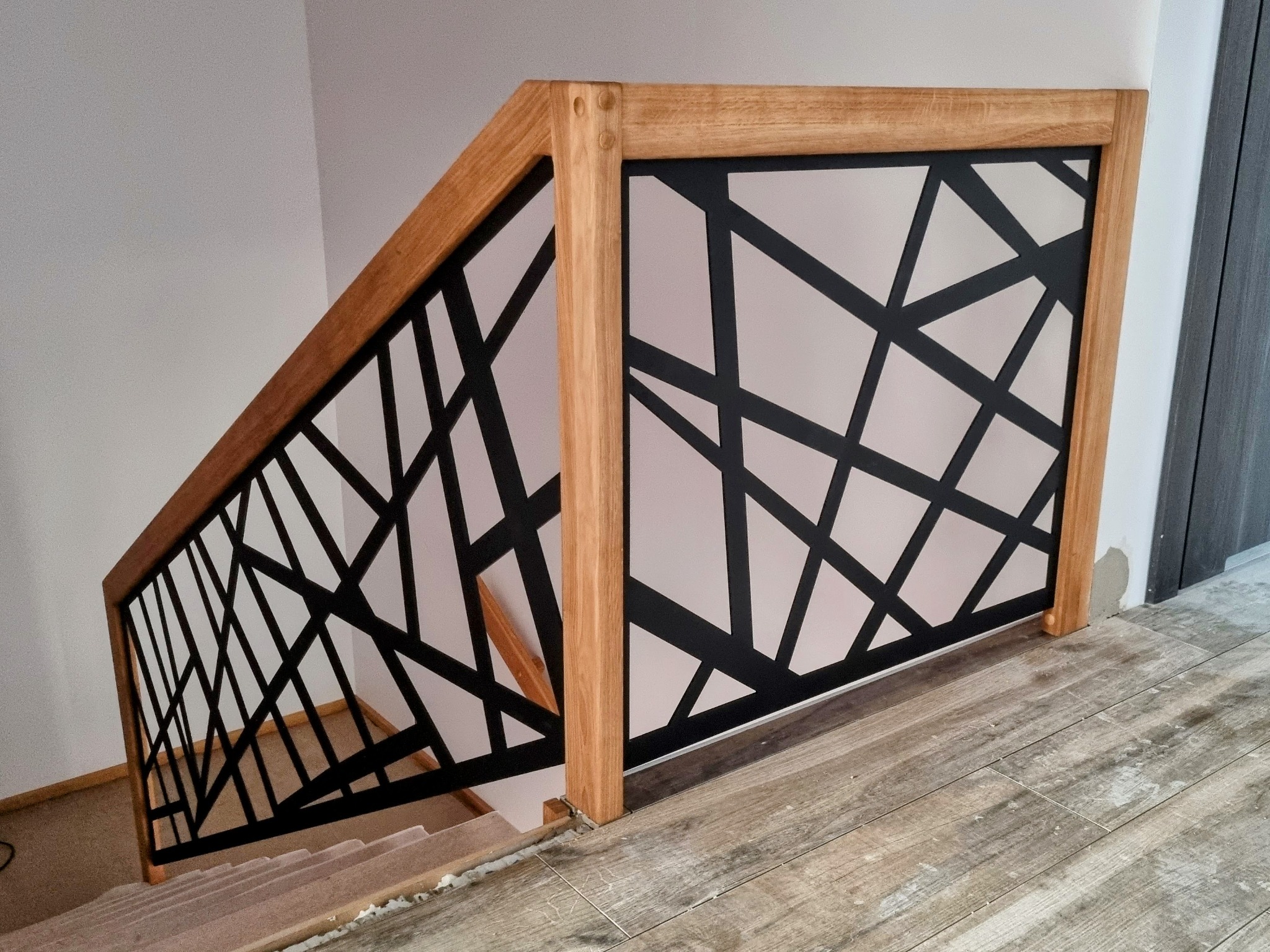 Nowoczesna balustrada schodowa z jasnego drewna z czarnym, geometrycznym wypełnieniem, widoczna z góry na tle białych ścian i drewnopodobnej podłogi.