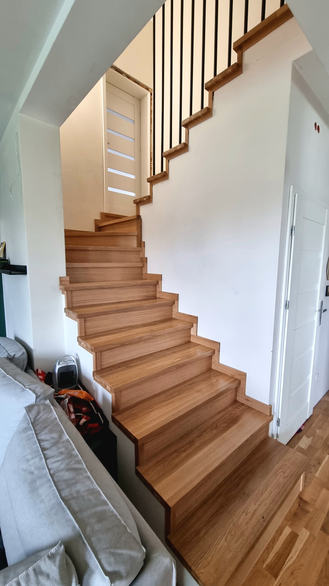 Nowoczesne, drewniane schody z czarnymi, metalowymi prętami balustrady, prowadzące w górę, widoczne z dołu obok sofy w jasnym salonie.