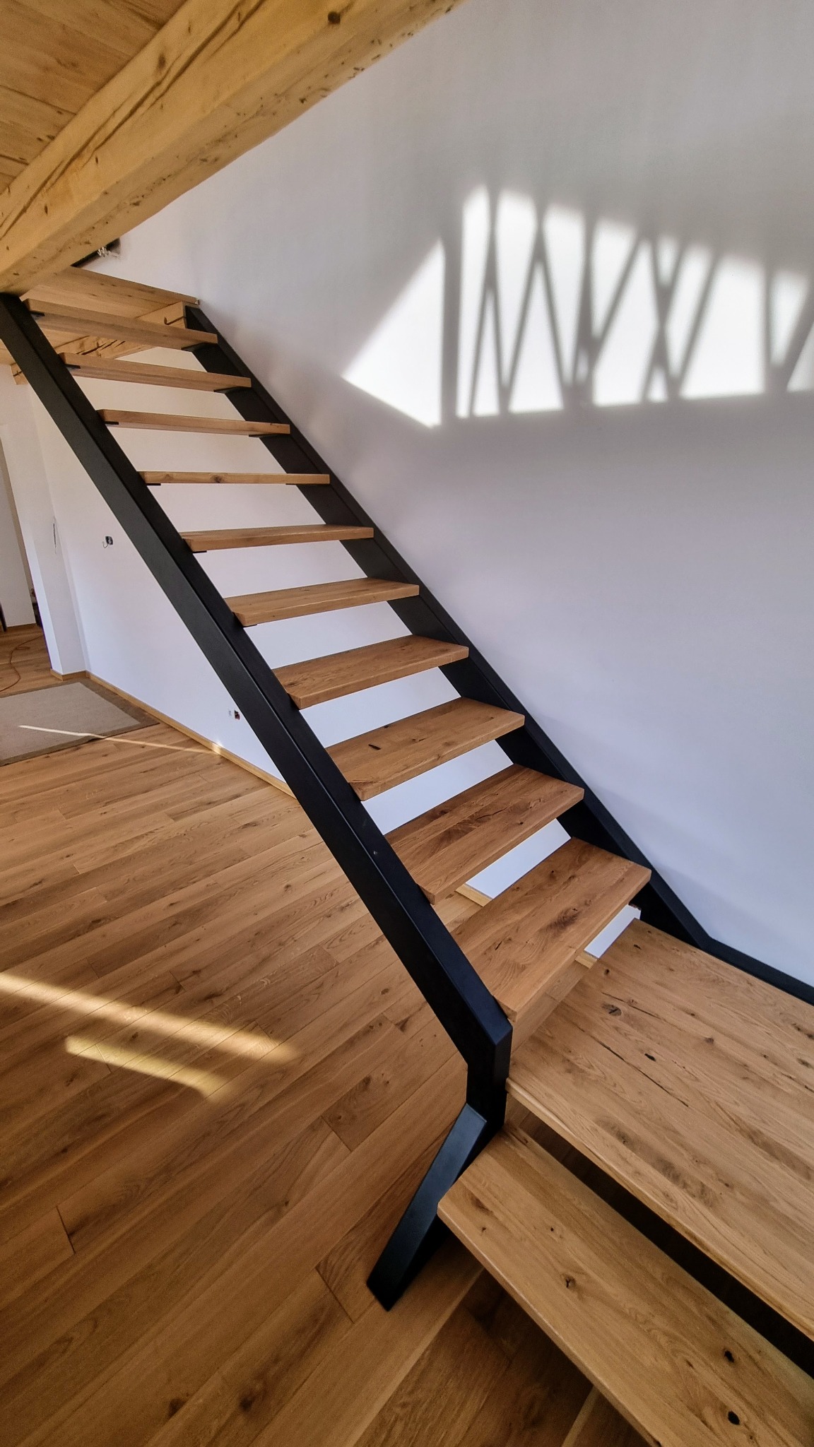 Nowoczesne schody z drewnianymi stopniami i czarną metalową konstrukcją, widoczne na tle białej ściany z cieniem, uzupełnione drewnianą podłogą i belką stropową.