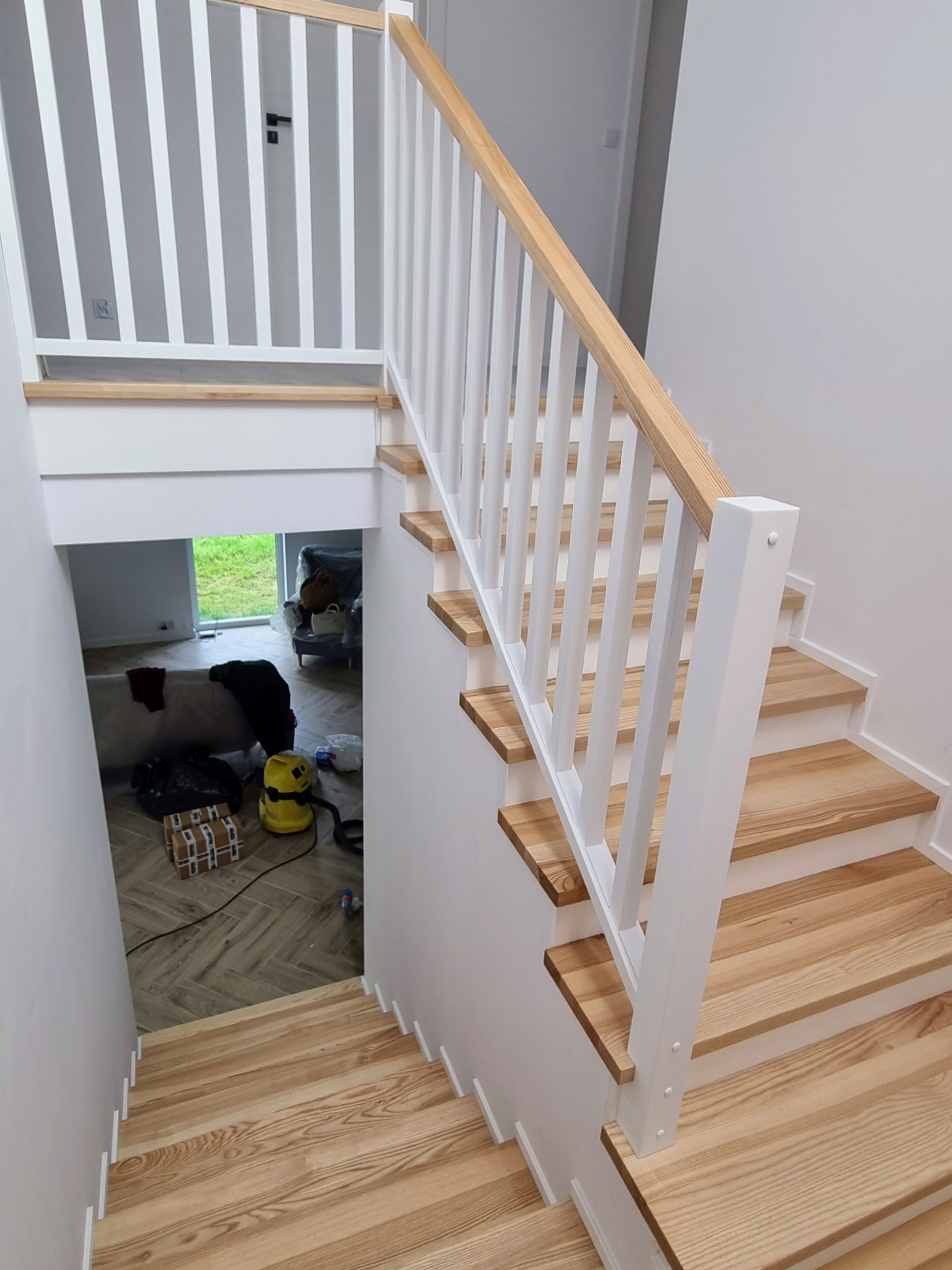 Jasne drewniane schody z białymi balustradami w nowoczesnym domu, widok z góry na dół, z widocznym pokojem i oknem w tle.