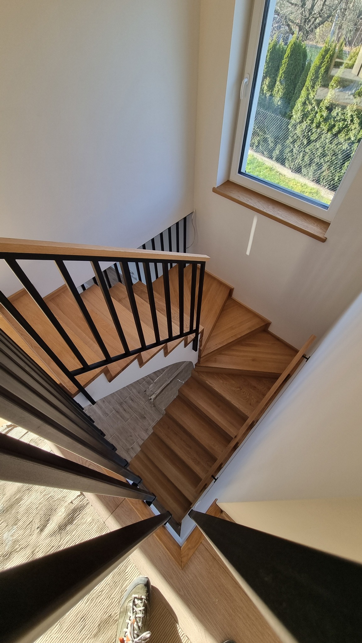 Spiralne stopnie z jasnego drewna z czarnymi metalowymi balustradami, widok z góry, z fragmentem buta w kadrze.