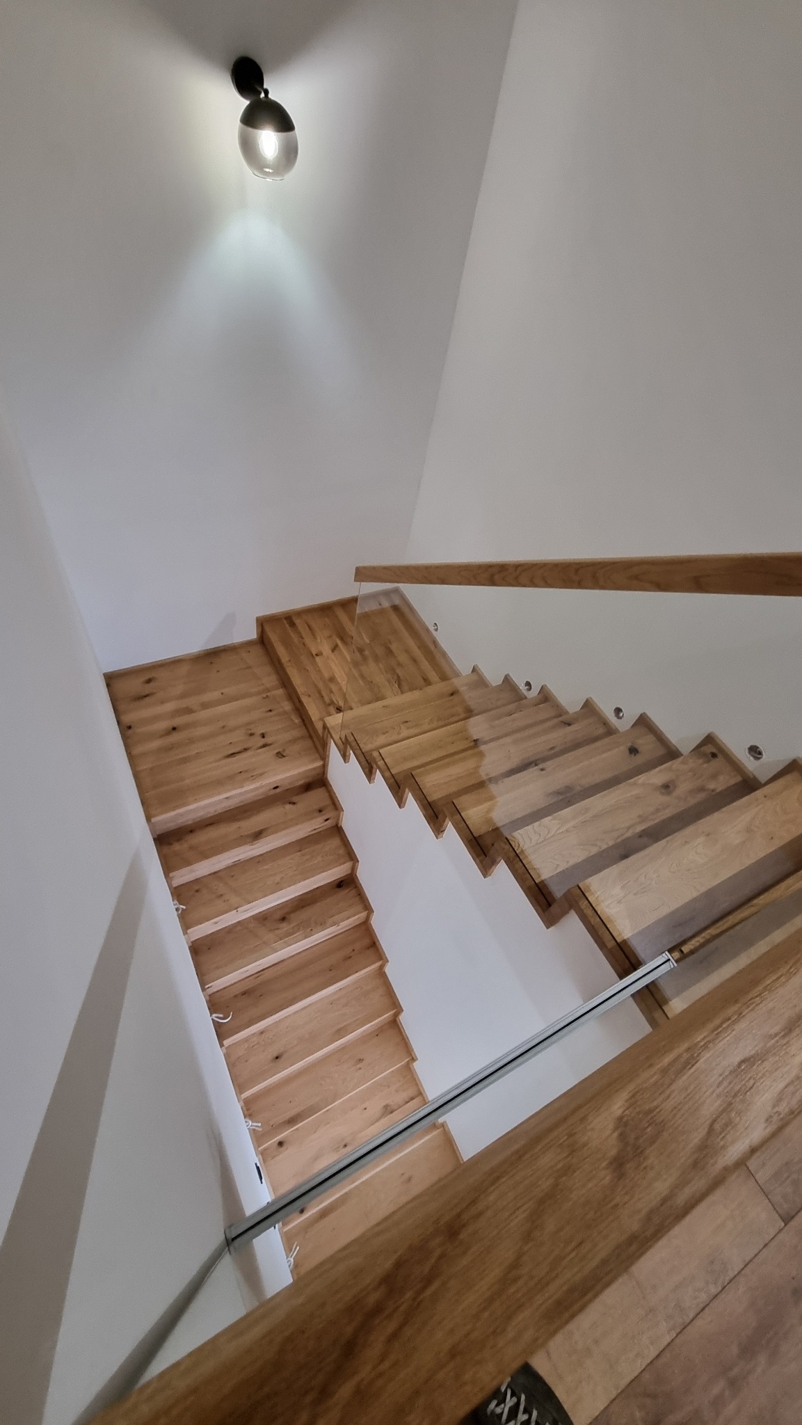 Jasne, dębowe stopnie schodów z przezroczystą balustradą szklaną, oświetlone punktowym światłem sufitowym, widok z góry.
