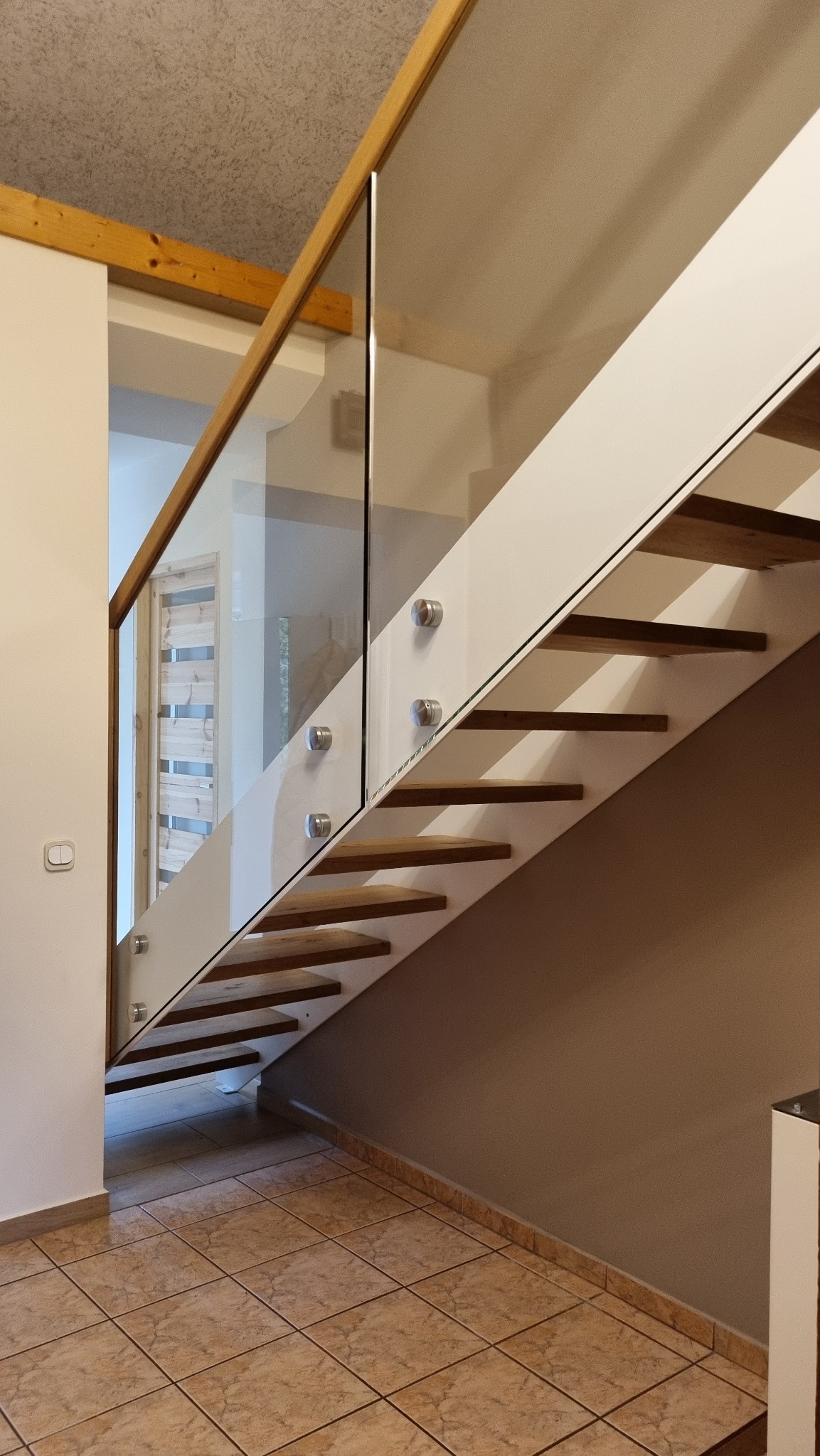 Nowoczesne schody z drewnianymi stopniami i szklaną balustradą mocowaną na metalowych uchwytach, widok z dołu, jasne wnętrze.