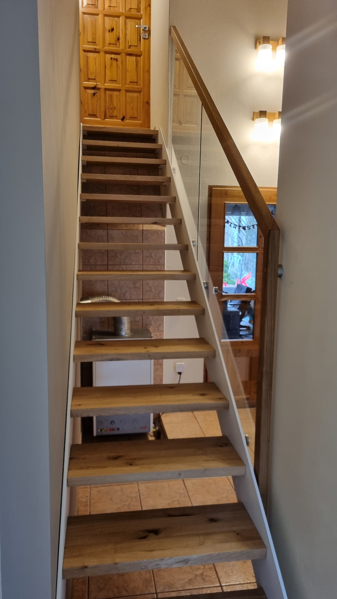 Wewnętrzne schody z drewnianymi stopniami i szklaną balustradą z drewnianą poręczą, prowadzące do drewnianych drzwi. Widoczny fragment grzejnika pod schodami.