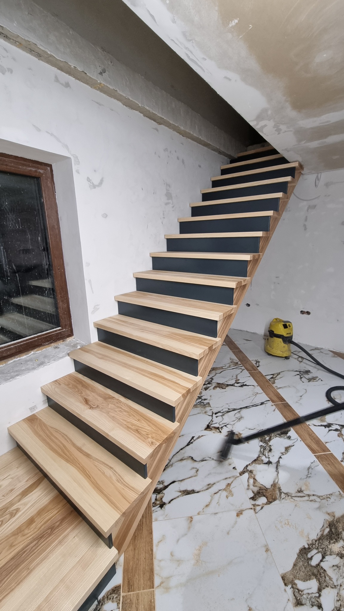 Wewnętrzne schody z jasnego drewna z ciemnymi podstopnicami, widoczne podczas montażu w surowym wnętrzu, z odkurzaczem przemysłowym w tle i marmurową podłogą.