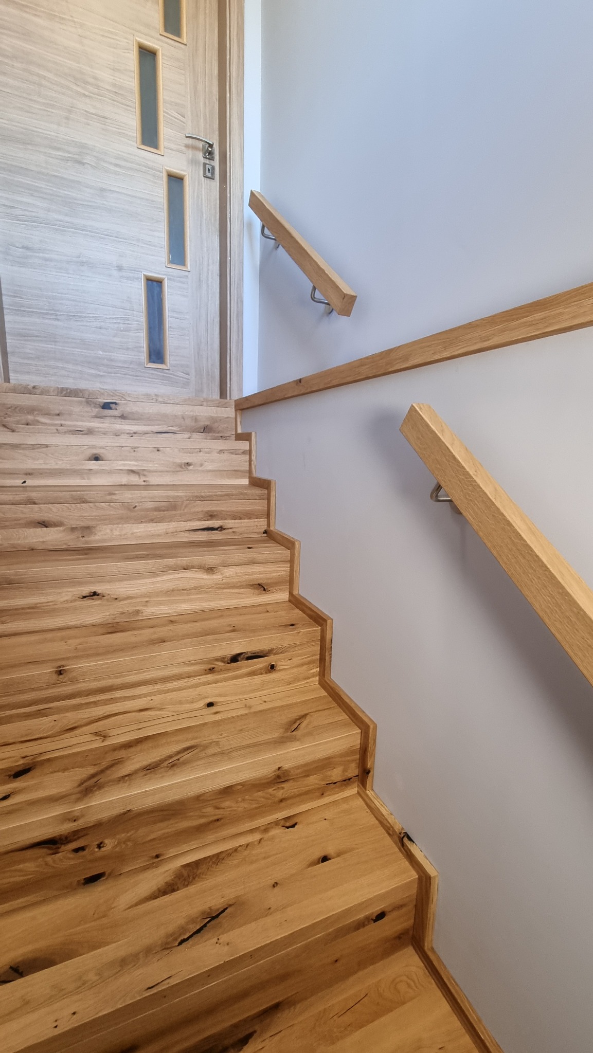 Jasne, drewniane schody z widocznymi sękami, z prostą, drewnianą poręczą mocowaną do ściany na metalowych wspornikach, prowadzące do drzwi z przeszkleniami.