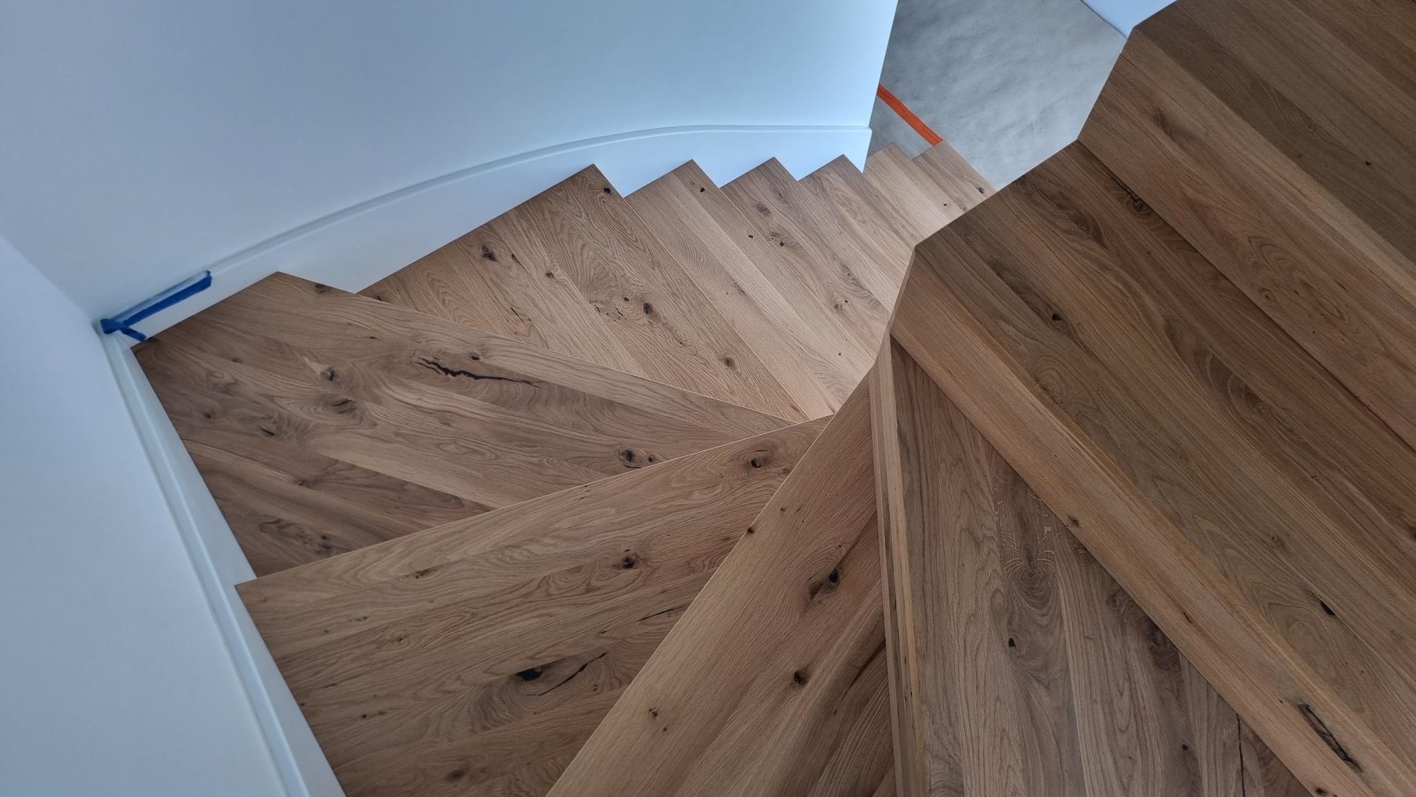 Widok z góry na drewniane schody z wyraźnym usłojeniem i naturalnymi sękami, zabezpieczone niebieską taśmą malarską przy białej ścianie.