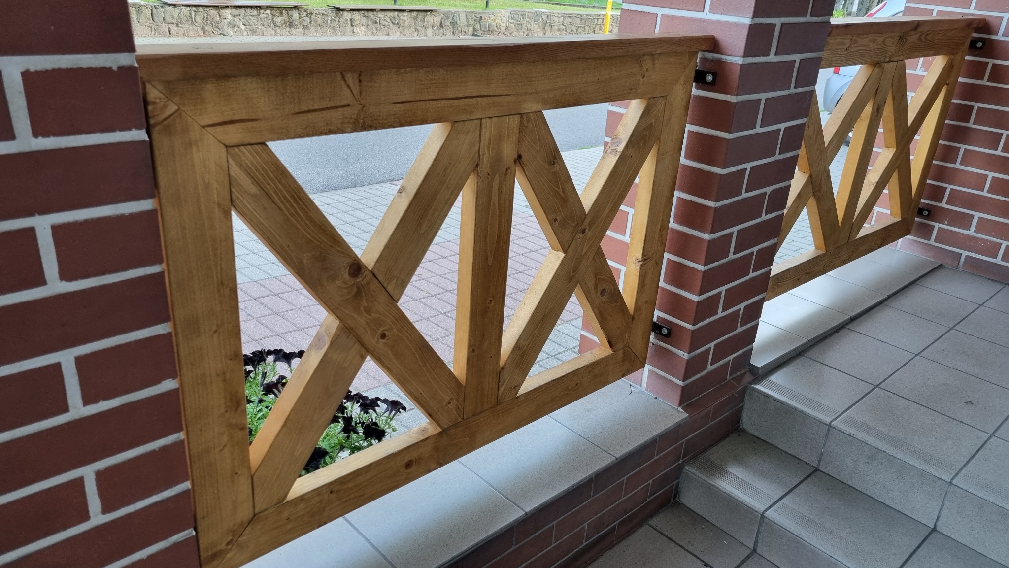 Fragment drewnianej balustrady z krzyżowym wzorem, mocowanej do ceglanych słupków, z widocznymi kwiatami w tle i fragmentem schodów.