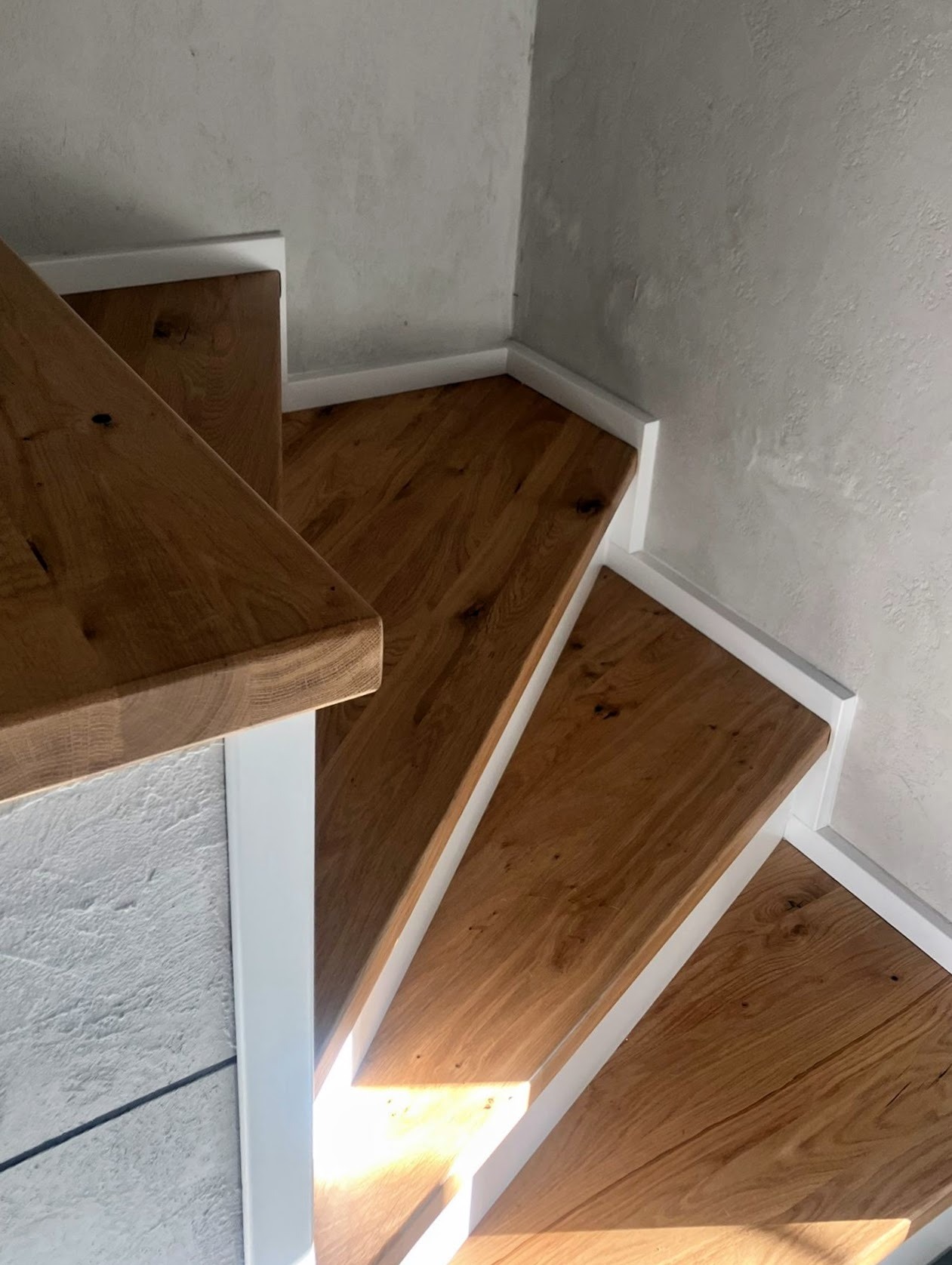 Widok z góry na dębowe stopnie schodów z białymi podstopnicami, prowadzące w górę, z widoczną fakturą drewna i szarą ścianą w tle.