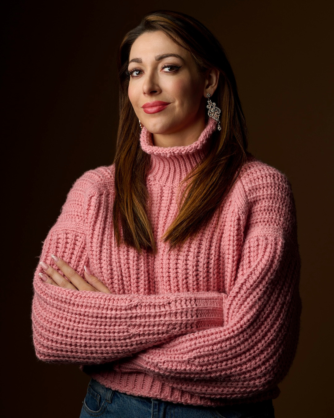 Portret kobiety z rękami skrzyżowanymi na piersiach, ubrana w różowy sweter z golfem i dżinsy, z dużymi, wiszącymi kolczykami na tle ciemnego, jednolitego tła.