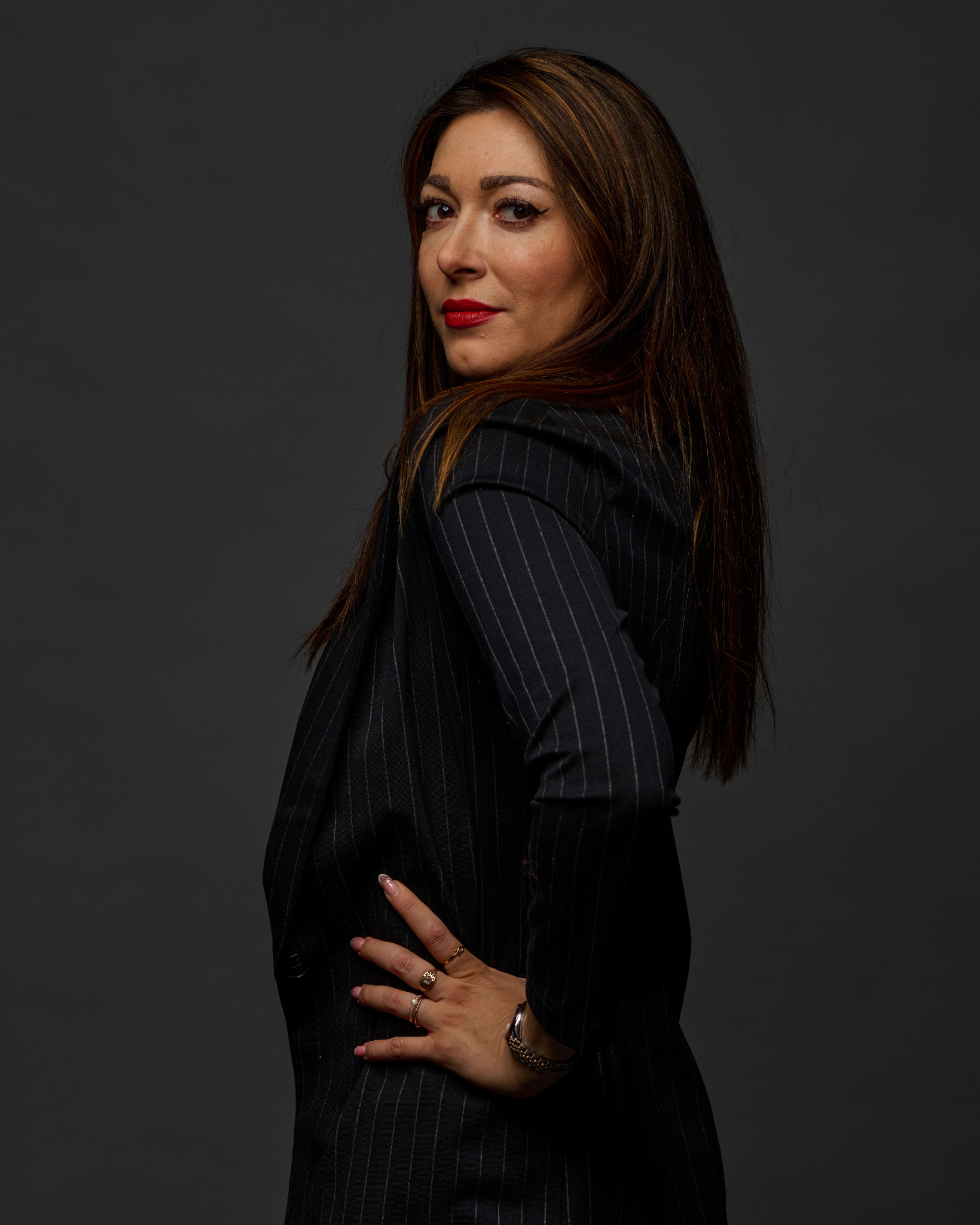 Portret kobiety w czarnym garniturze w prążki, odwróconej przez ramię, z czerwonymi ustami i biżuterią na dłoni, na jednolitym szarym tle.