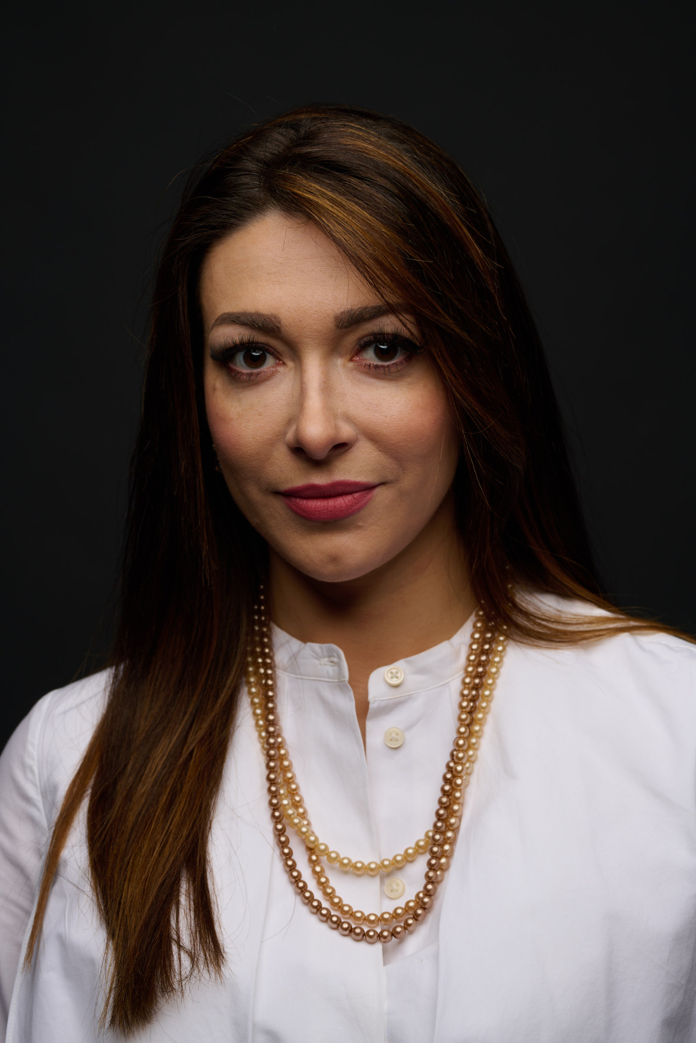 Portret kobiety z brązowymi włosami i perłowym naszyjnikiem na ciemnoszarym tle, ubrana w białą koszulę.