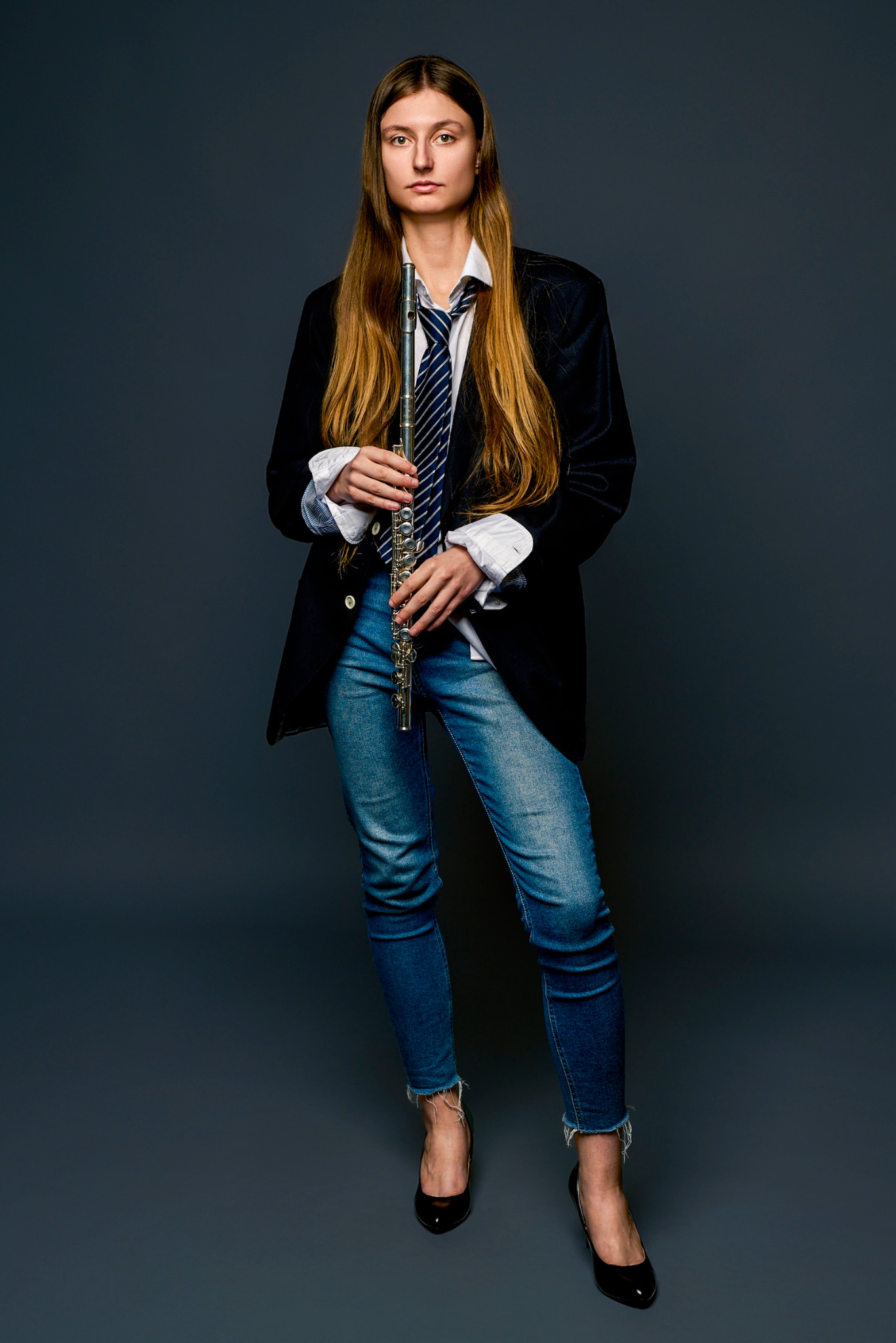 Portret kobiety z długimi blond włosami, w rozpiętej koszuli, marynarce i dżinsach, trzymającej flet poprzeczny na jednolitym szarym tle, pozującej do zdjęcia.
