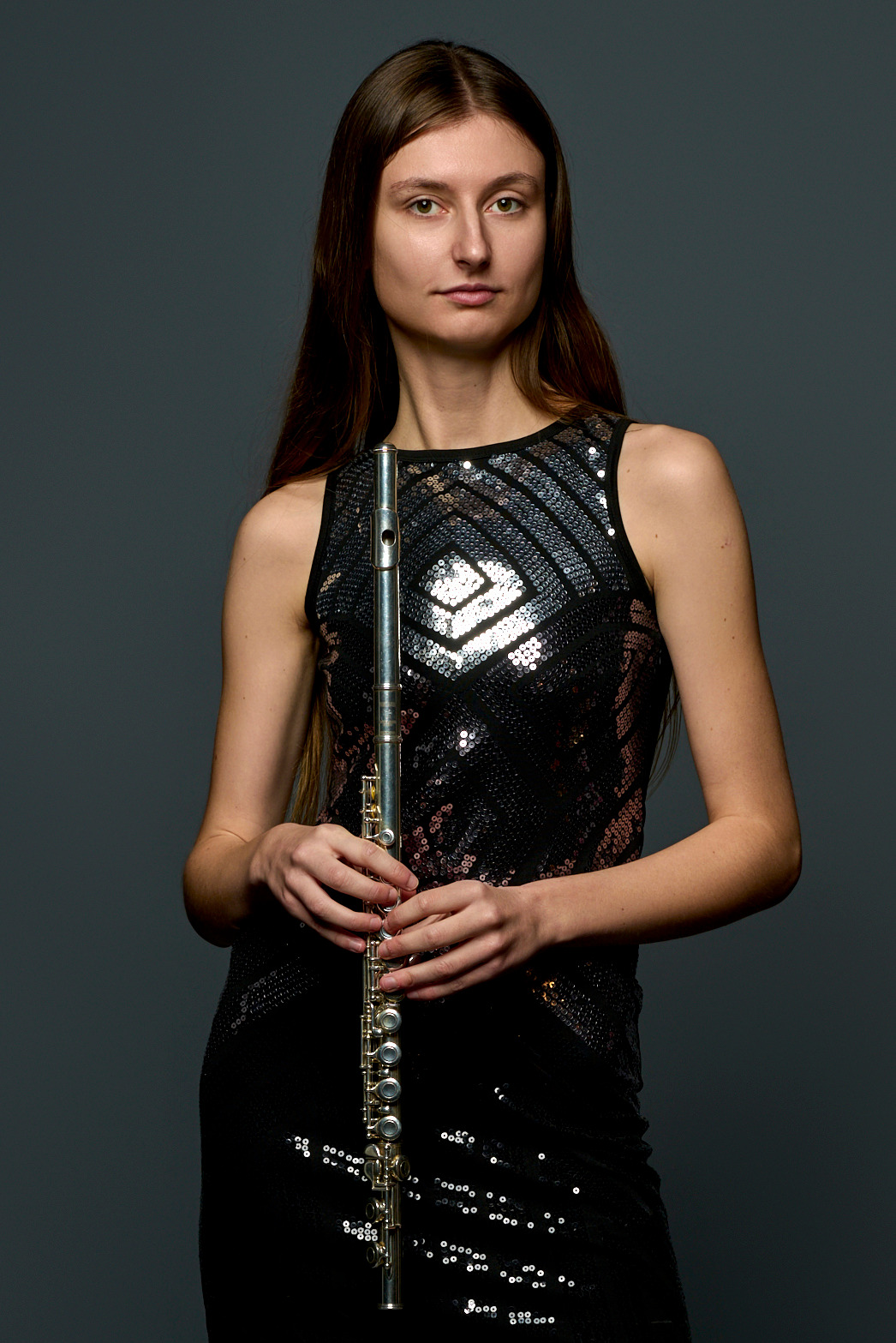 Portret kobiety z długimi włosami, trzymającej flet poprzeczny na szarym tle, ubrana w czarną sukienkę z cekinami.