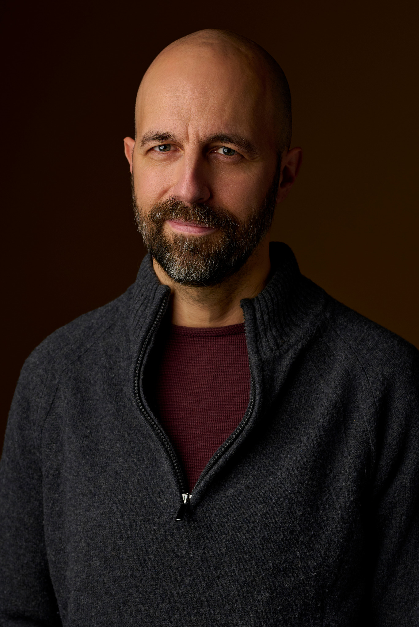 Portret mężczyzny z brodą w ciemnoszarym swetrze na tle ciemnobrązowego tła, oświetlony studyjnym światłem.