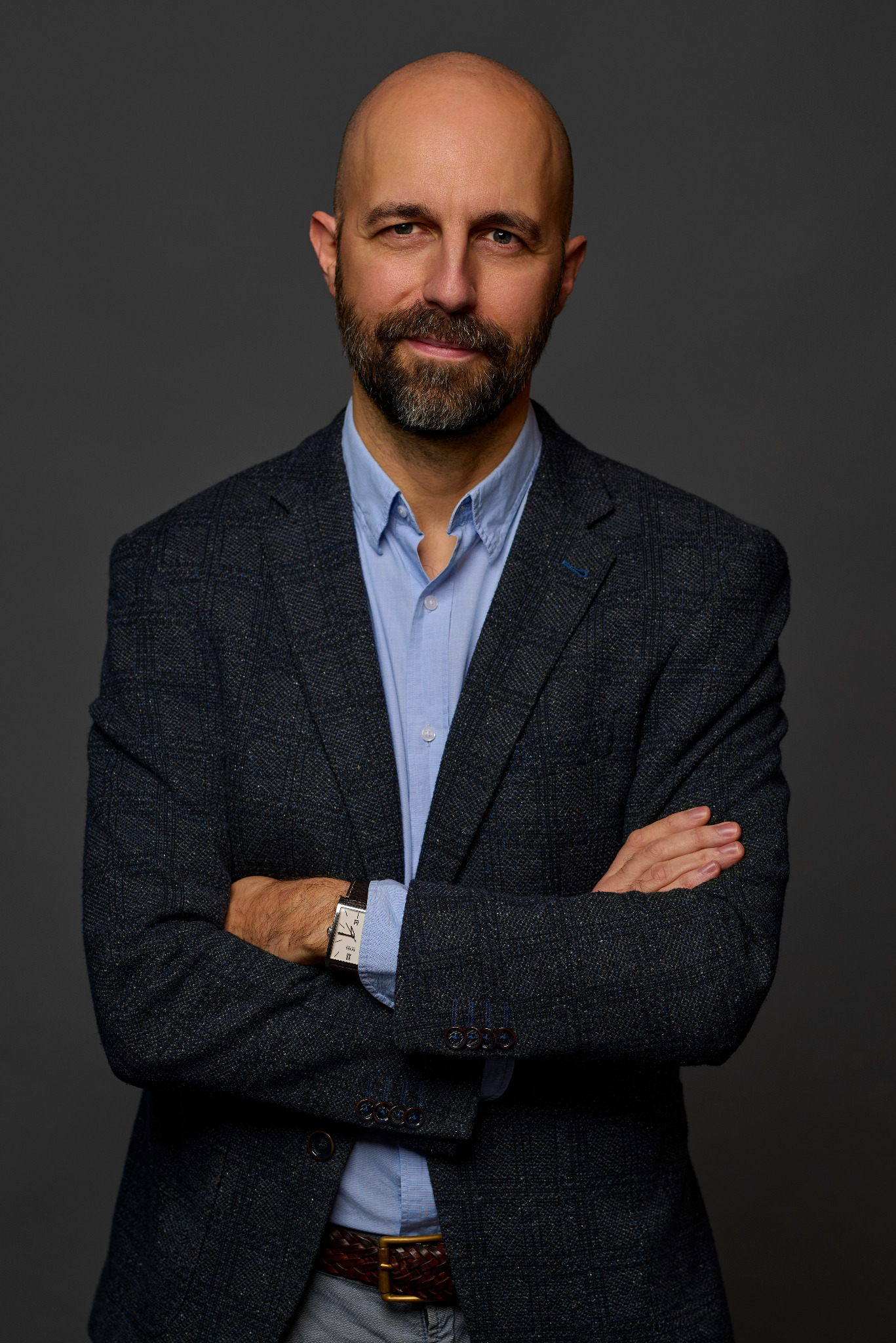 Portret mężczyzny z brodą w niebieskiej koszuli i tweedowej marynarce, z założonymi rękami, na szarym tle. Widoczny zegarek na skórzanym pasku i skórzany pleciony pasek.