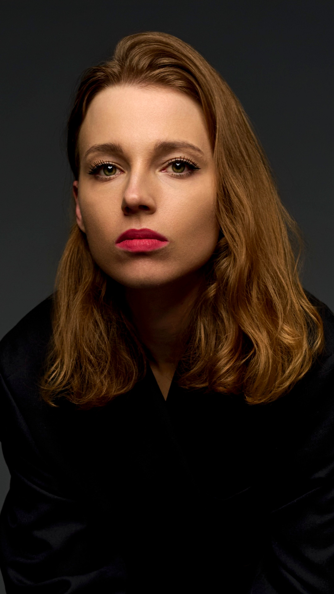 Portret kobiety z zielonymi oczami i czerwonymi ustami, w czarnej marynarce na ciemnoszarym tle, profesjonalne oświetlenie.