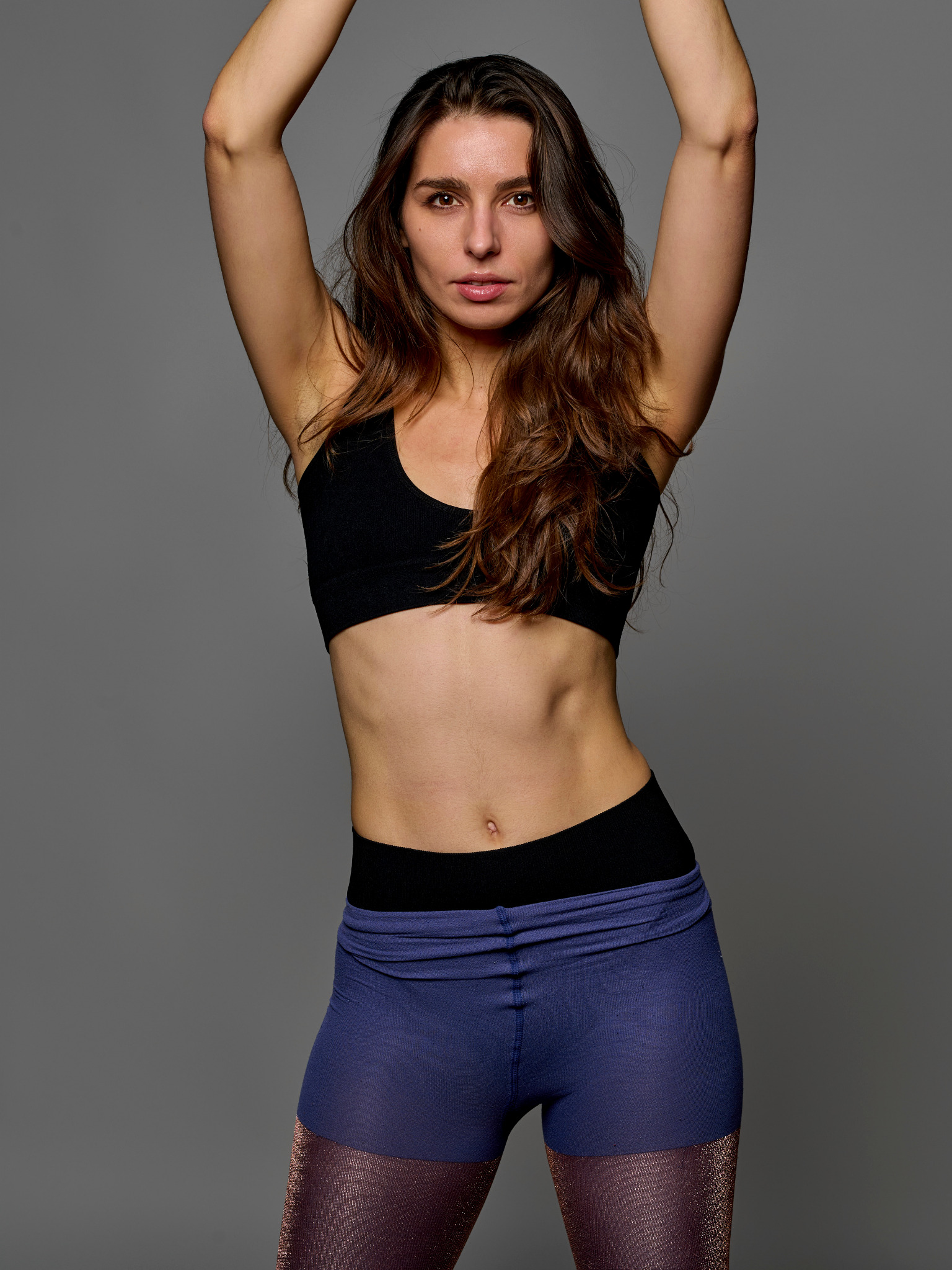 Portret kobiety w sportowym stroju na szarym tle, z rękami uniesionymi nad głową, eksponujący sylwetkę.