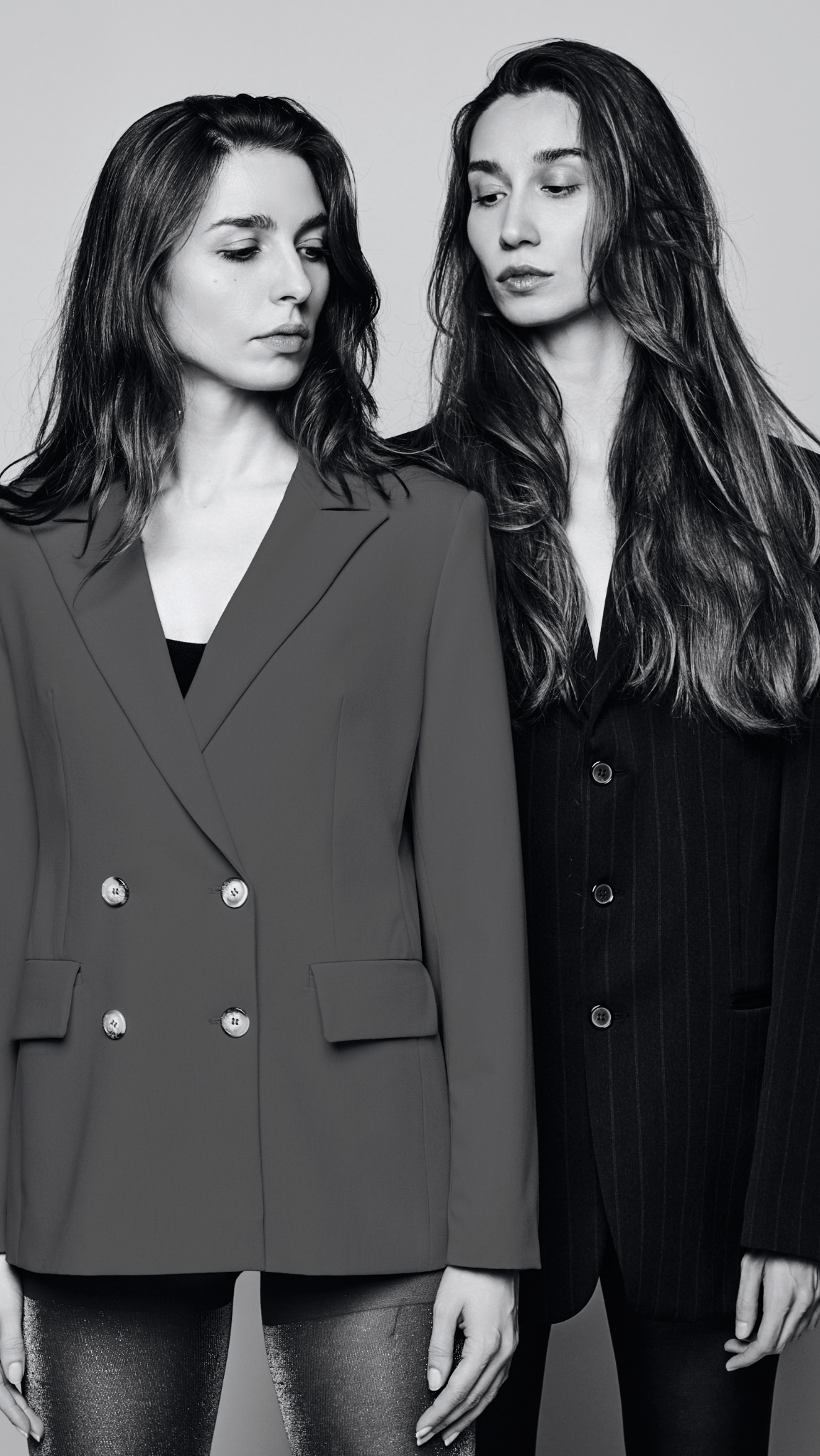 Dwie kobiety pozują w eleganckich marynarkach na monochromatycznym tle, jedna w czerwonej dwurzędowej marynarce z dużymi guzikami, druga w czarnej marynarce w prążki, obie z długimi, falowanymi...