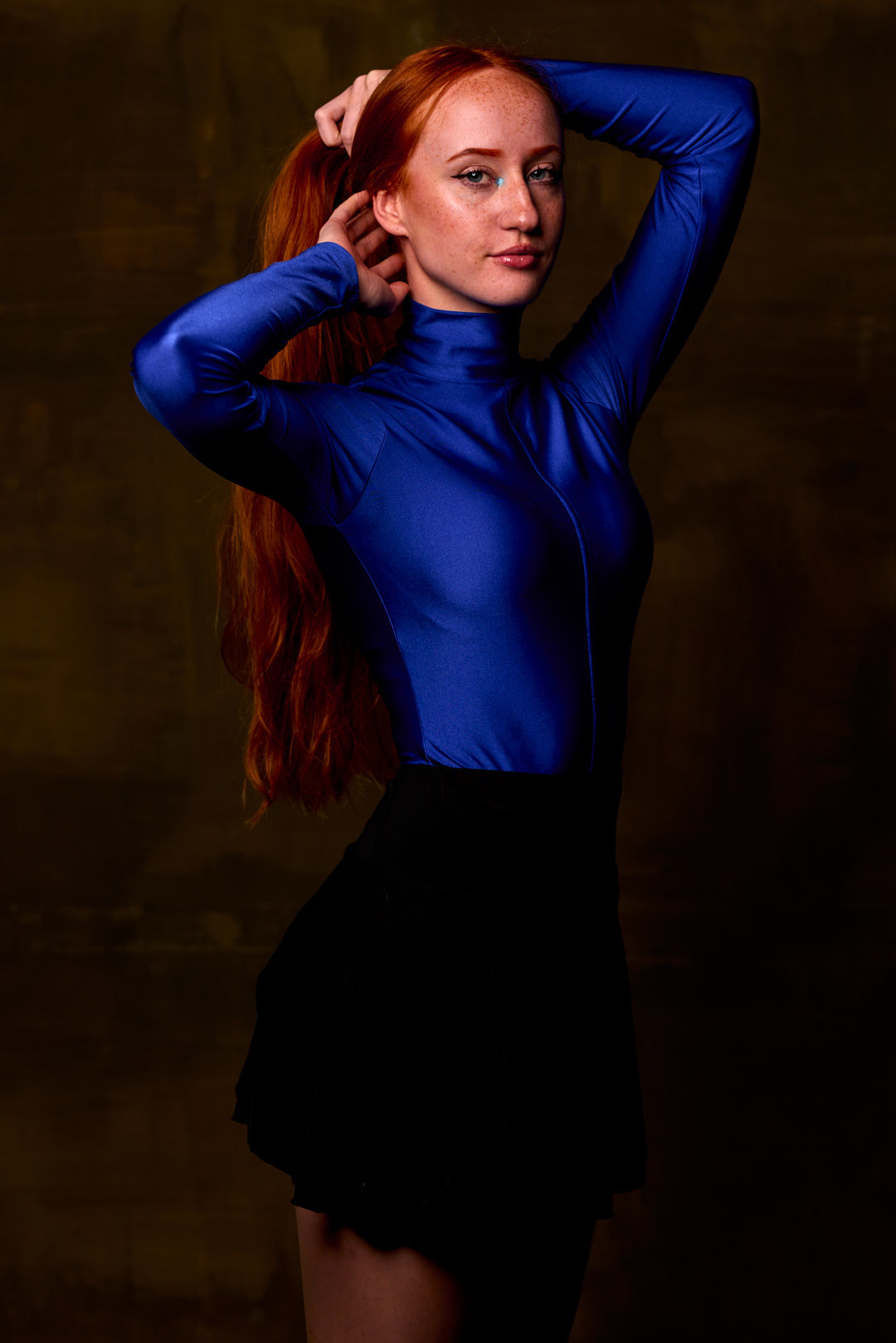 Portret kobiety z rudymi włosami, w niebieskim body i czarnej spódnicy, pozującej na tle ciemnego, teksturowanego tła, z subtelnym makijażem oczu.