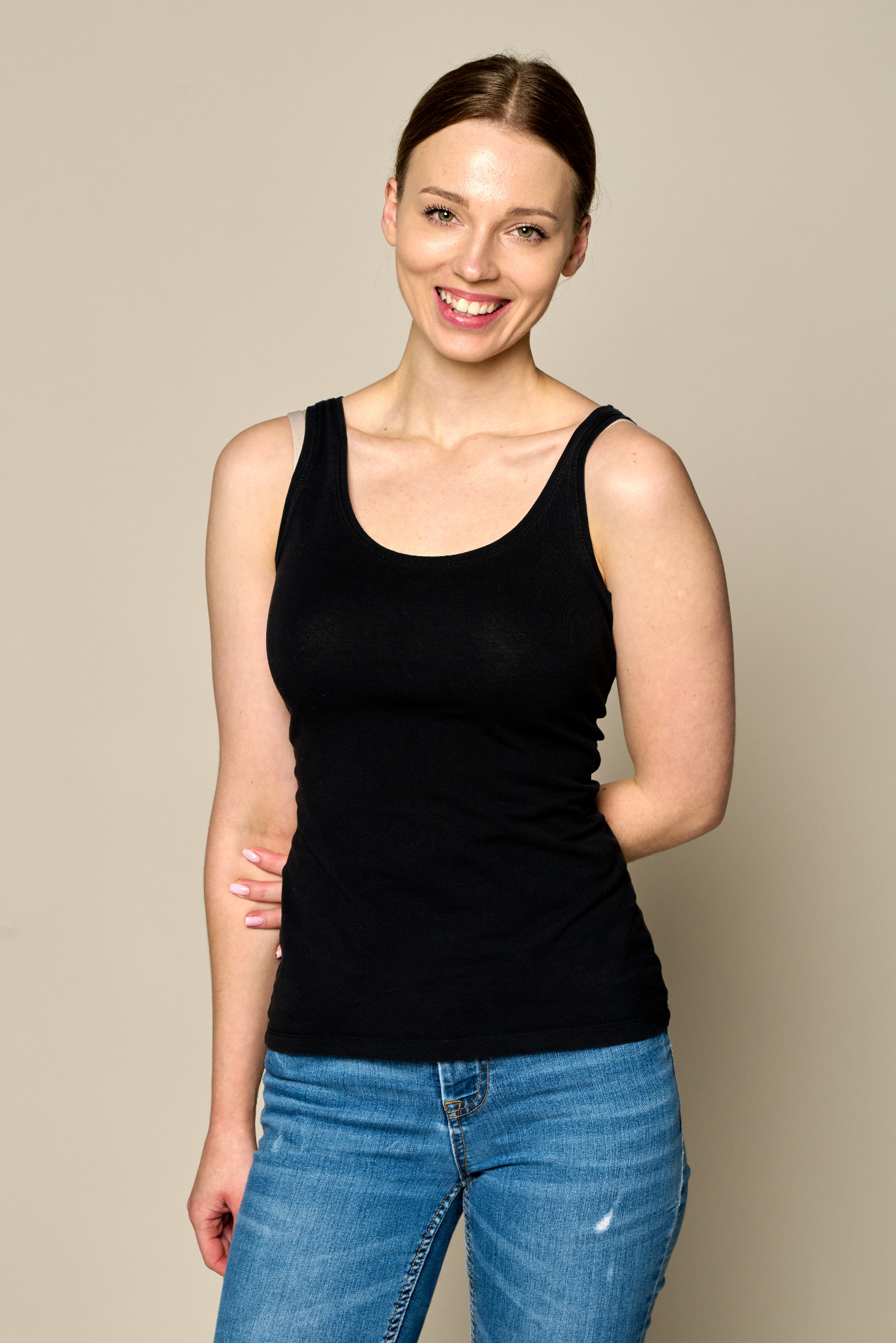 Uśmiechnięta kobieta w czarnej koszulce na ramiączkach i dżinsach, stojąca na jasnym tle, z rękami założonymi za plecami i delikatnym makijażem.
