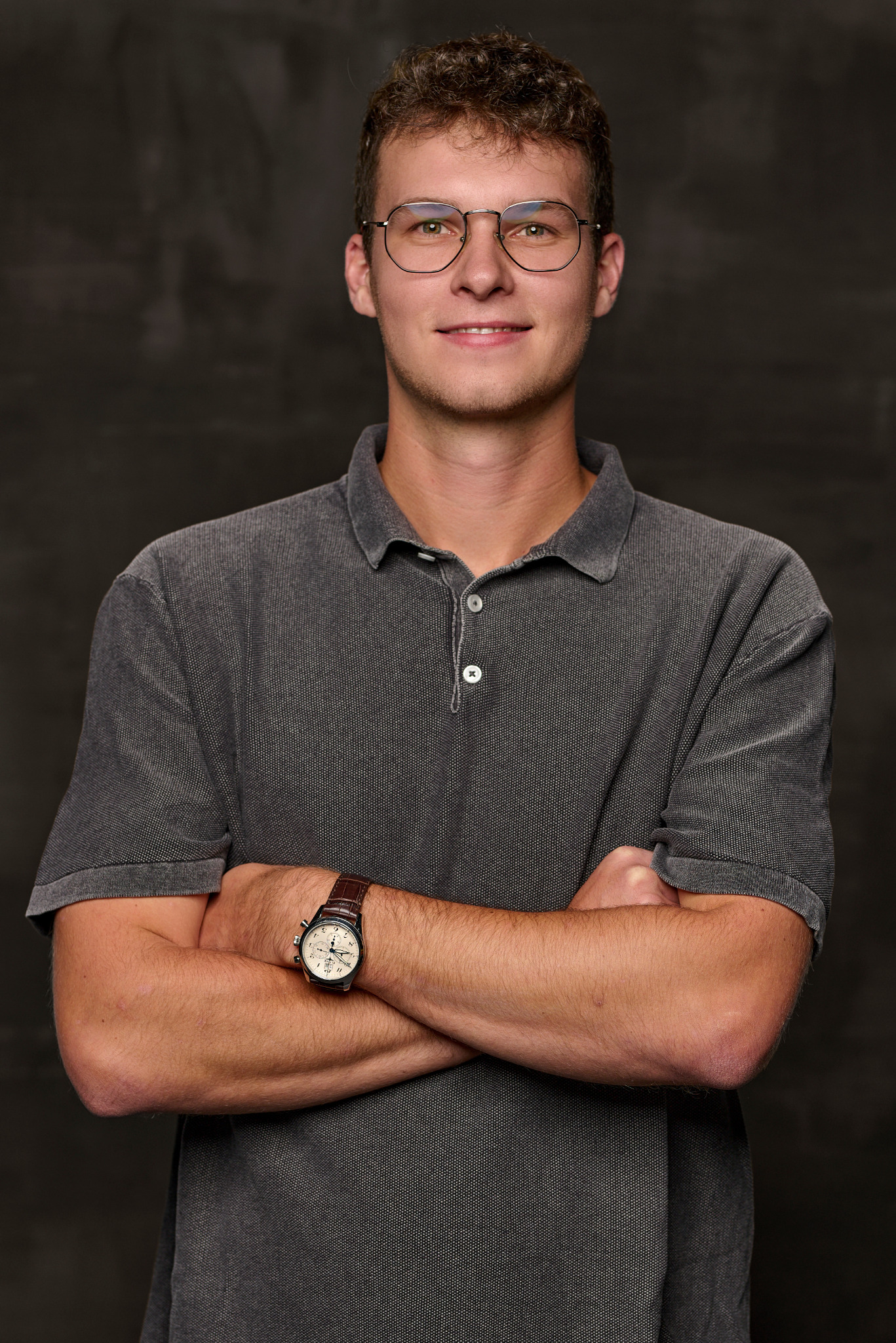 Portret młodego mężczyzny w okularach z założonymi rękami, widoczny zegarek na skórzanym pasku, szara koszulka polo, ciemne tło.