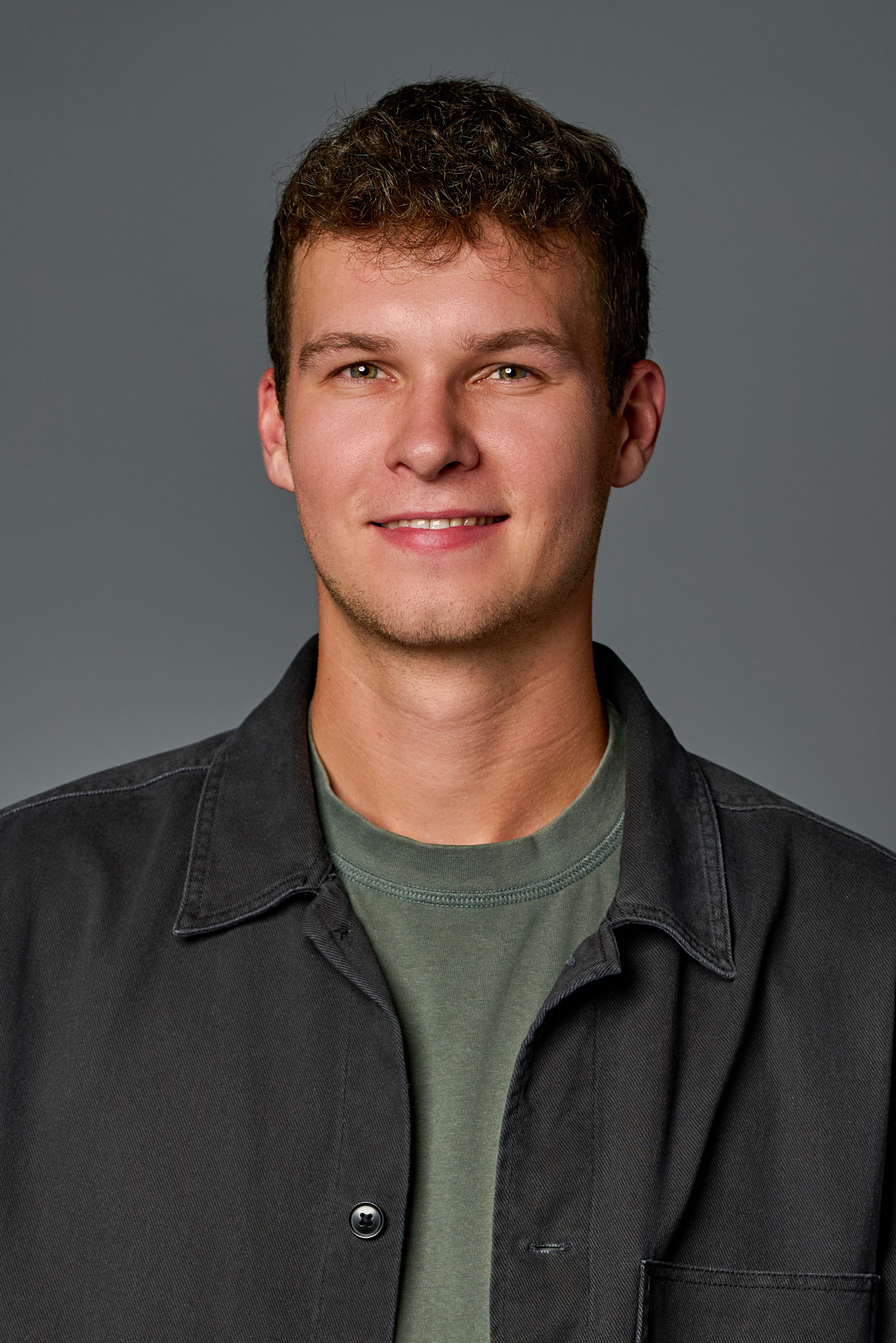 Portret uśmiechniętego młodego mężczyzny z brązowymi włosami na szarym tle, ubrany w zieloną koszulkę i ciemnoszarą koszulę z kołnierzykiem.