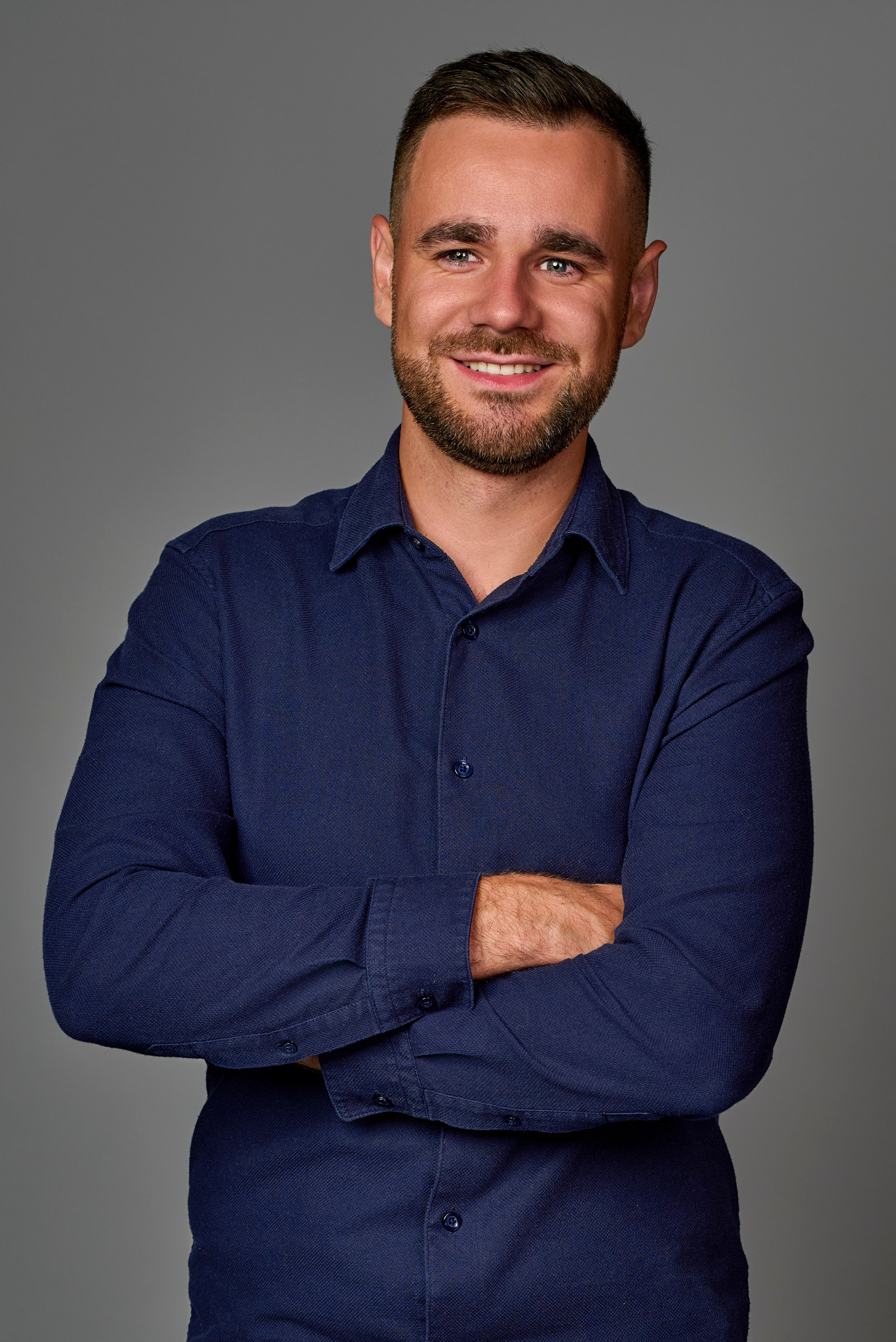 Uśmiechnięty mężczyzna z brodą, ubrany w granatową koszulę, pozuje z założonymi rękami na szarym tle.