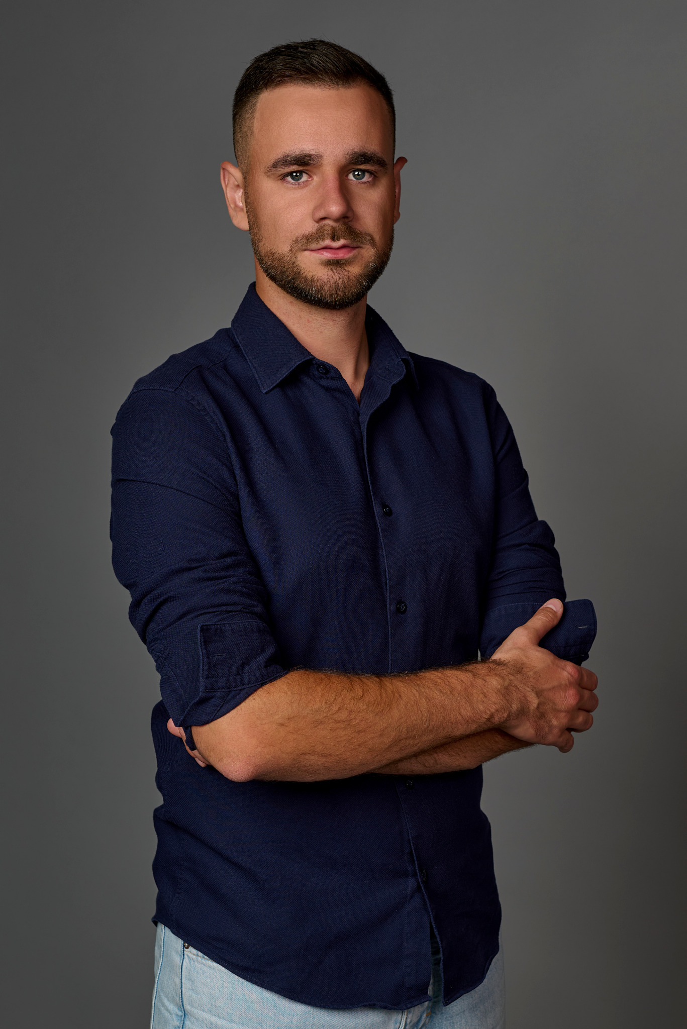 Mężczyzna z brodą, ubrany w granatową koszulę, pozuje na szarym tle, z rękami skrzyżowanymi na piersiach.