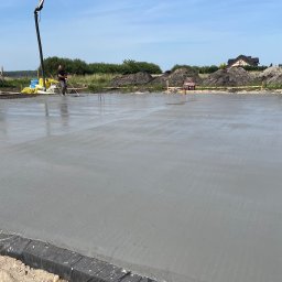 KP Budownictwo - Wylewka fundamentowa z betonu zbrojonego, wyrównywana przez dwóch pracowników z użyciem długich łat, z betoniarką i stertami materiałów w tle.