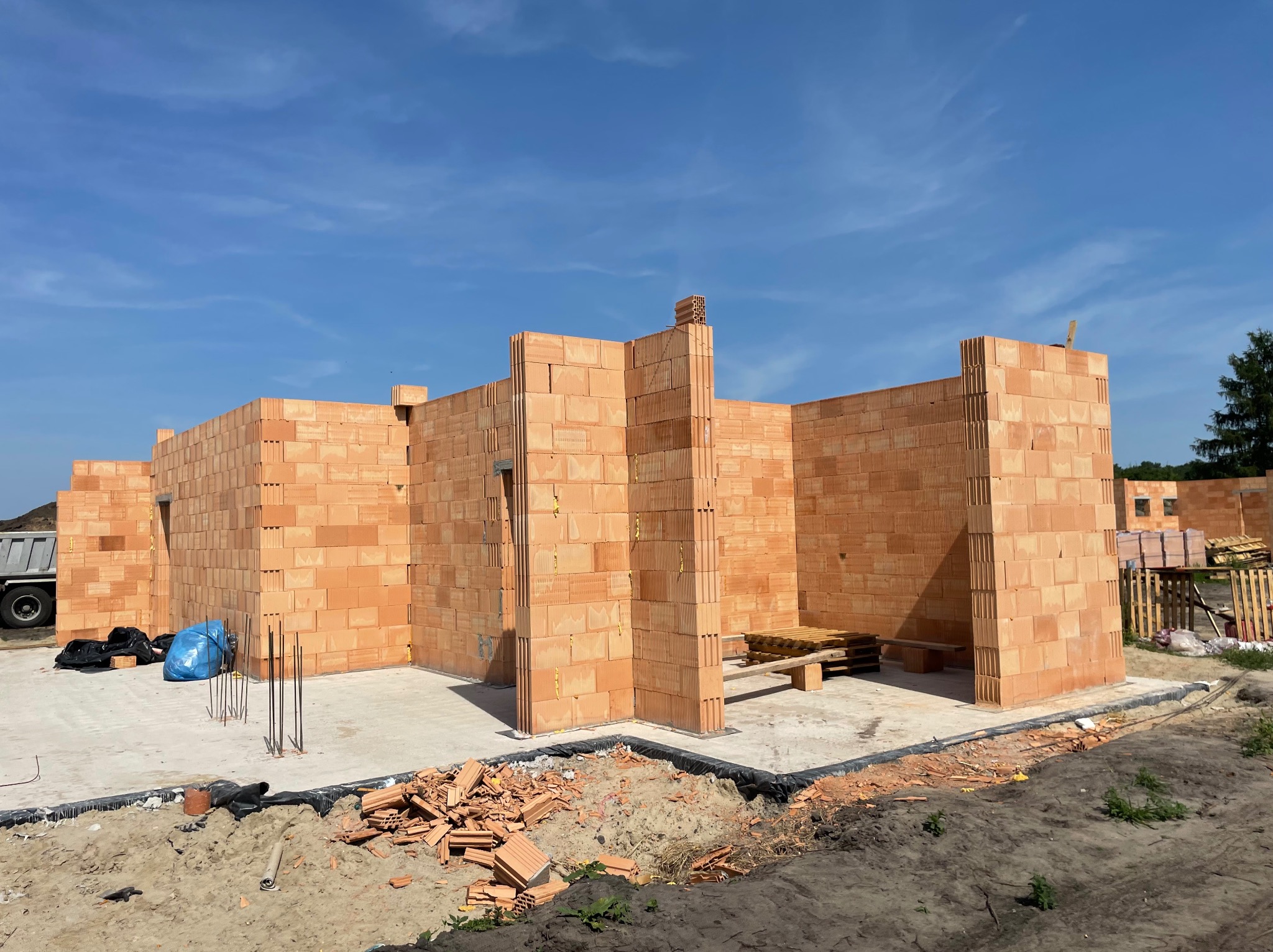 Wznoszenie ścian parterowego domu z pustaków ceramicznych, widoczne wystające pręty zbrojeniowe, gruz ceglany i palety wewnątrz budynku, na zewnątrz zaparkowana ciężarówka.