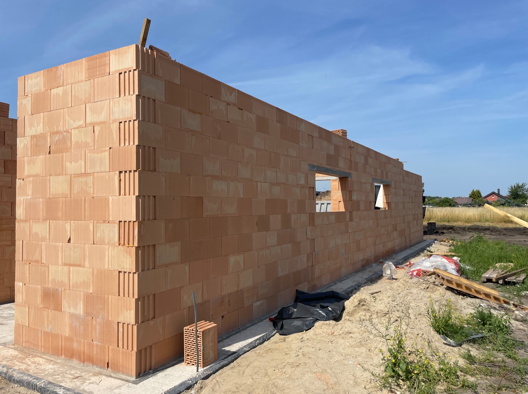 Wznoszenie ściany z czerwonej ceramiki poryzowanej, widoczne otwory okienne, na zewnątrz plac budowy z piaskiem i resztkami materiałów budowlanych.