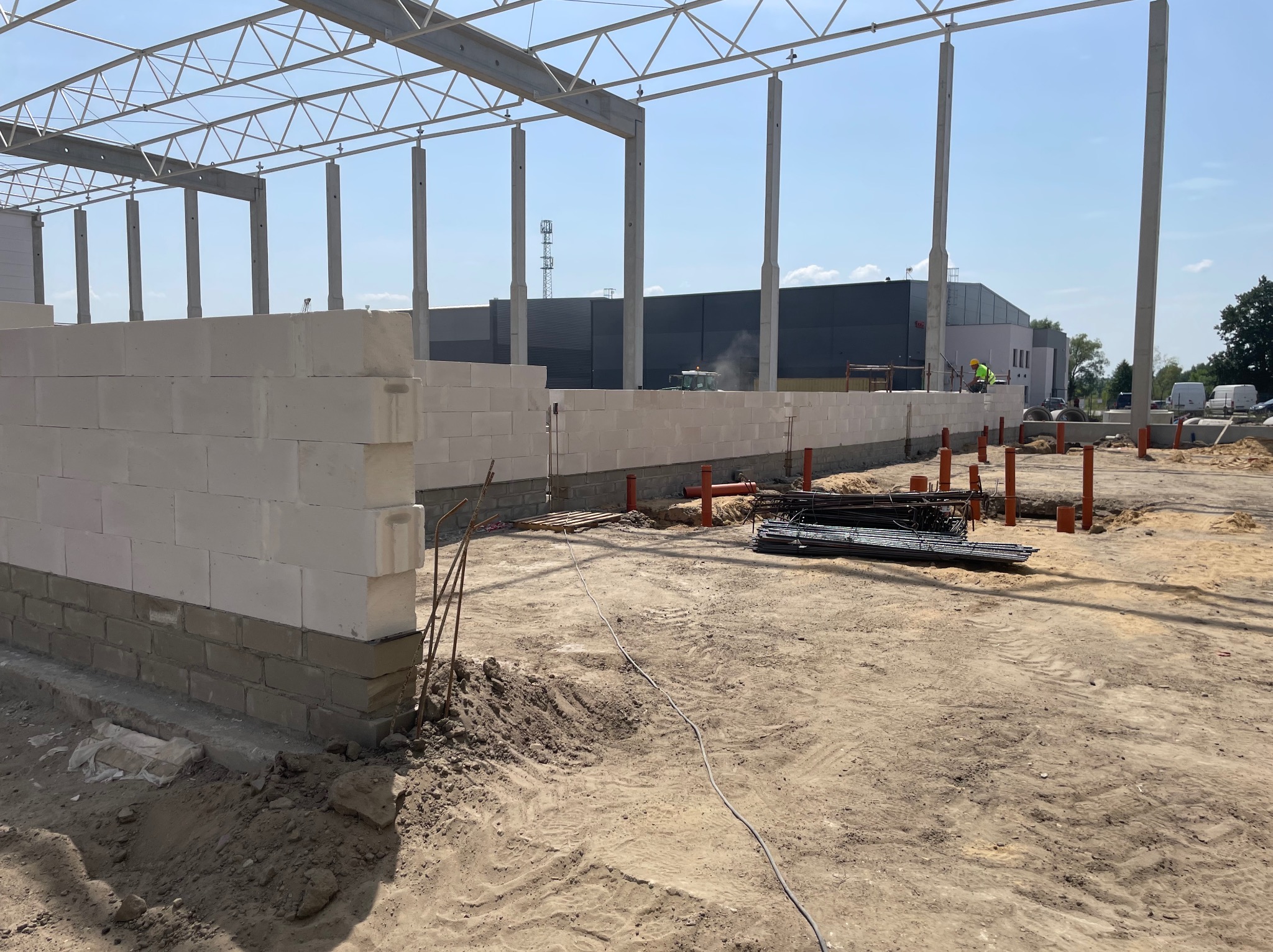Budowa hali: wznoszenie ścian z bloczków na fundamencie, widoczne stalowe elementy konstrukcyjne dachu, pomarańczowe rury instalacyjne i sterta prętów zbrojeniowych na piaszczystym terenie.