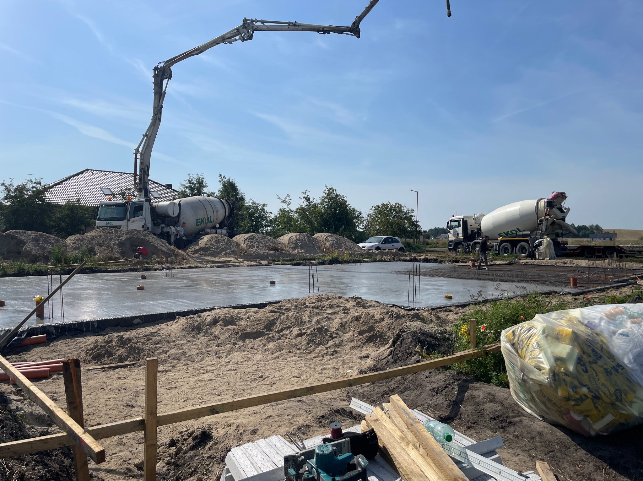 Wylewanie betonu na przygotowany fundament z wystającymi prętami zbrojeniowymi, widoczne dwie betoniarki firmy EKOL i pompa do betonu na tle błękitnego nieba.