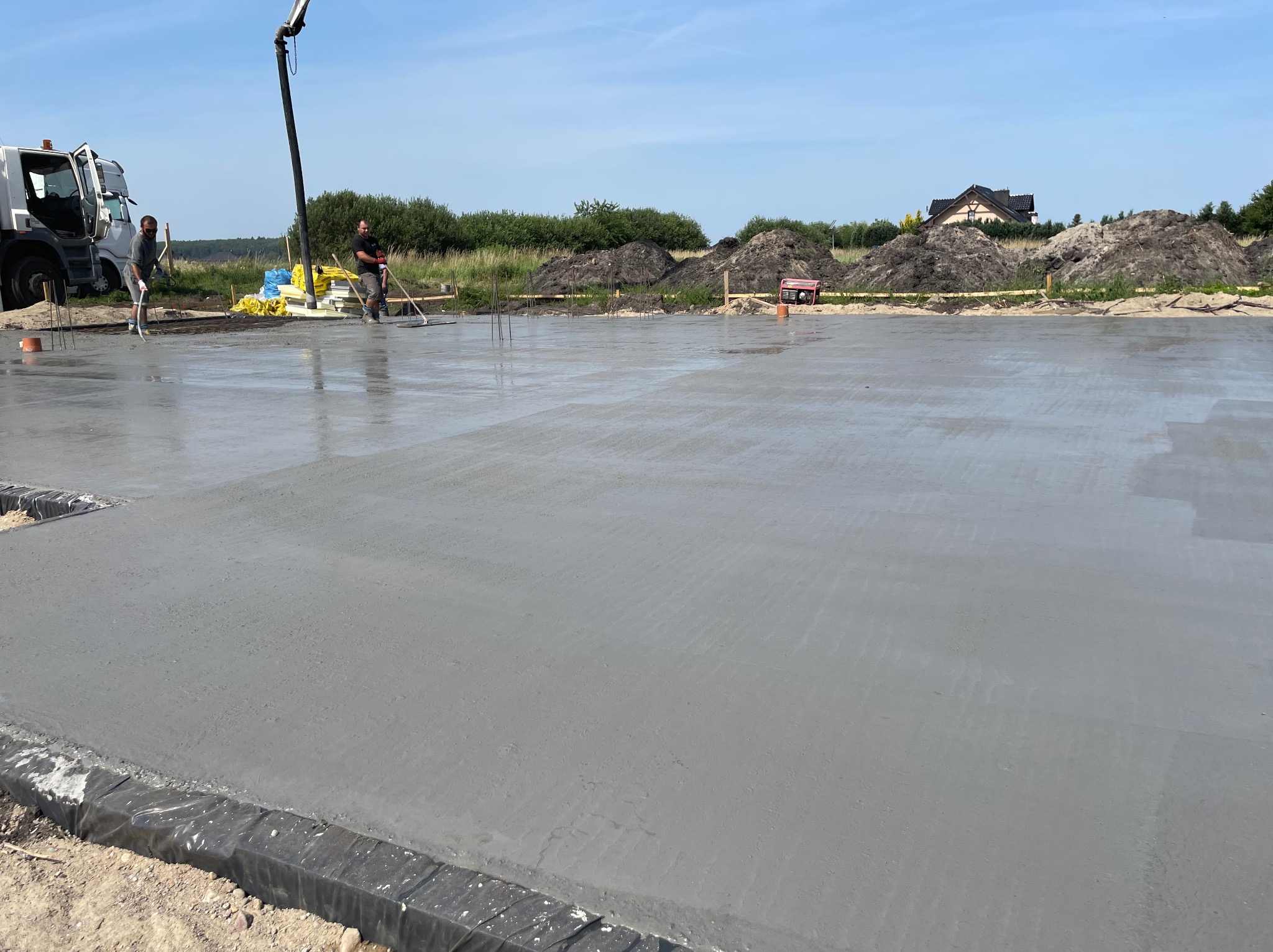 Wylewka fundamentowa z betonu zbrojonego, wyrównywana przez dwóch pracowników z użyciem długich łat, z betoniarką i stertami materiałów w tle.