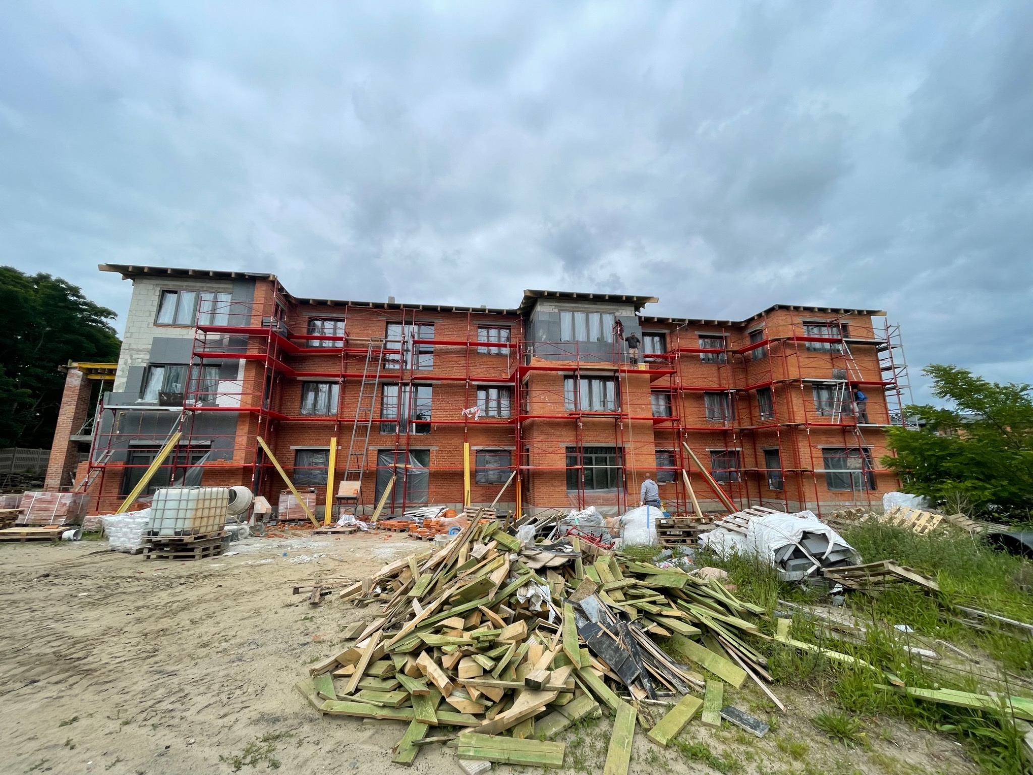 Wielopiętrowy budynek w trakcie budowy z czerwonej cegły, otoczony rusztowaniami, z widocznym stosem drewnianych odpadów i materiałów budowlanych na pierwszym planie.