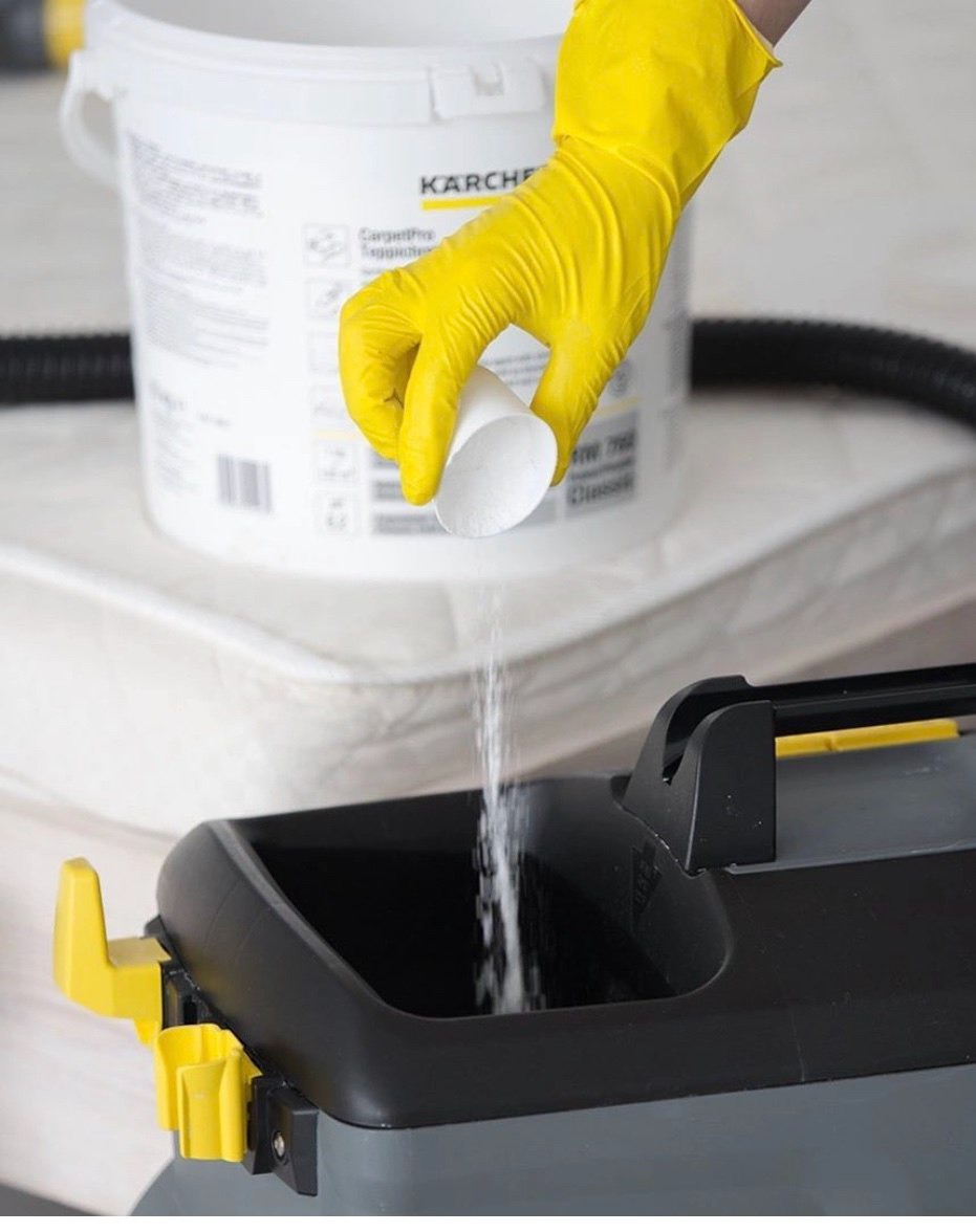 Wsypywanie proszku do czyszczenia z miarki do zbiornika odkurzacza piorącego Karcher, widoczne żółte rękawice ochronne i wiadro z napisem CarpetPro, w tle materac.