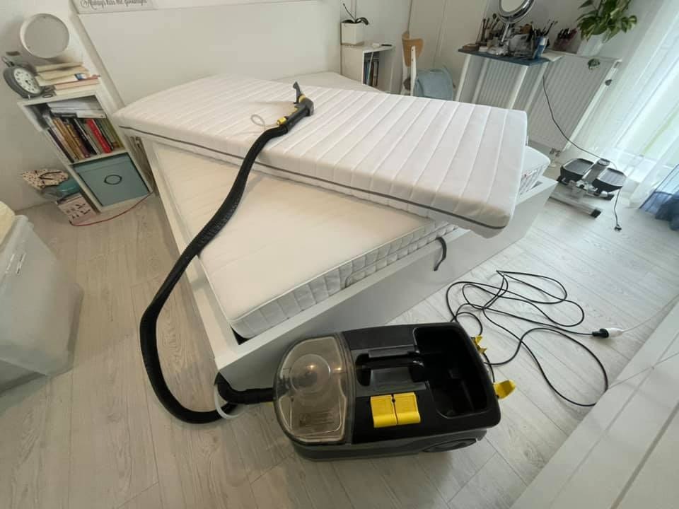 Czyszczenie materaca parowym odkurzaczem z żółtymi akcentami, widoczne dwa materace na łóżku, jeden na drugim, w tle książki i dekoracje.