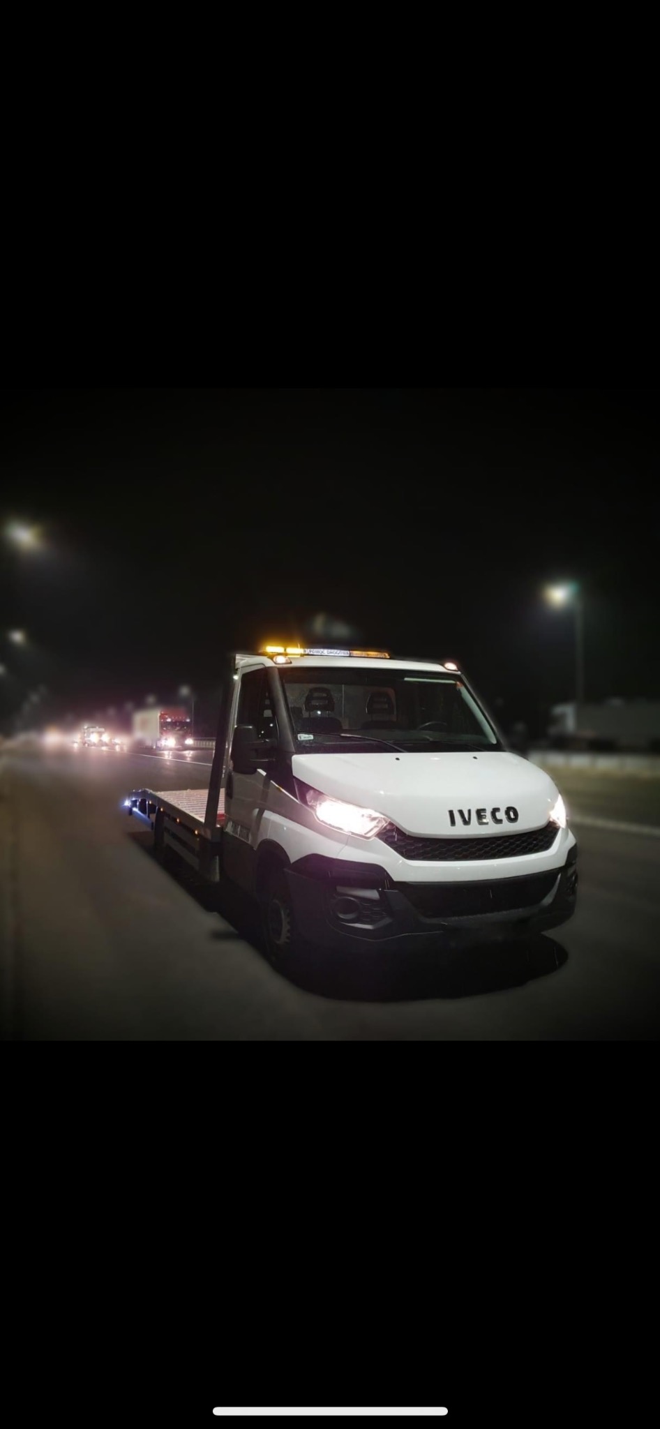 Biała laweta Iveco z włączonymi światłami ostrzegawczymi, widok z przodu, nocna sceneria z rozmazanymi światłami innych pojazdów w tle.