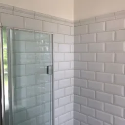 Białe, prostokątne płytki ułożone w jodełkę na ścianie kabiny prysznicowej z szklanymi drzwiami i srebrnymi okuciami.