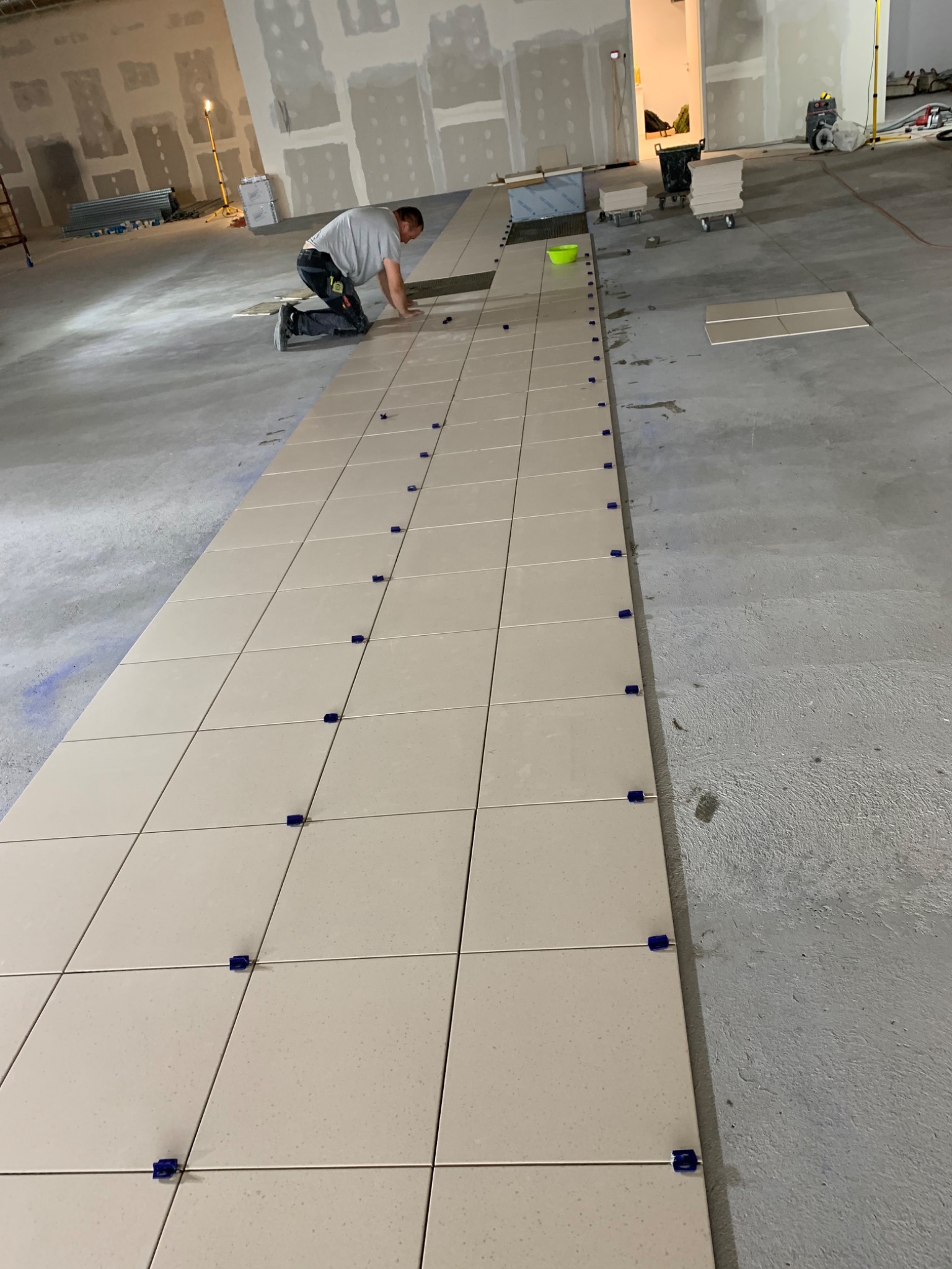 Układanie jasnych, kwadratowych płytek na podłodze w dużym pomieszczeniu. Glazurnik na kolanach wyrównuje płytki za pomocą niebieskich krzyżyków dystansowych. W tle widoczne przygotowane płyty...