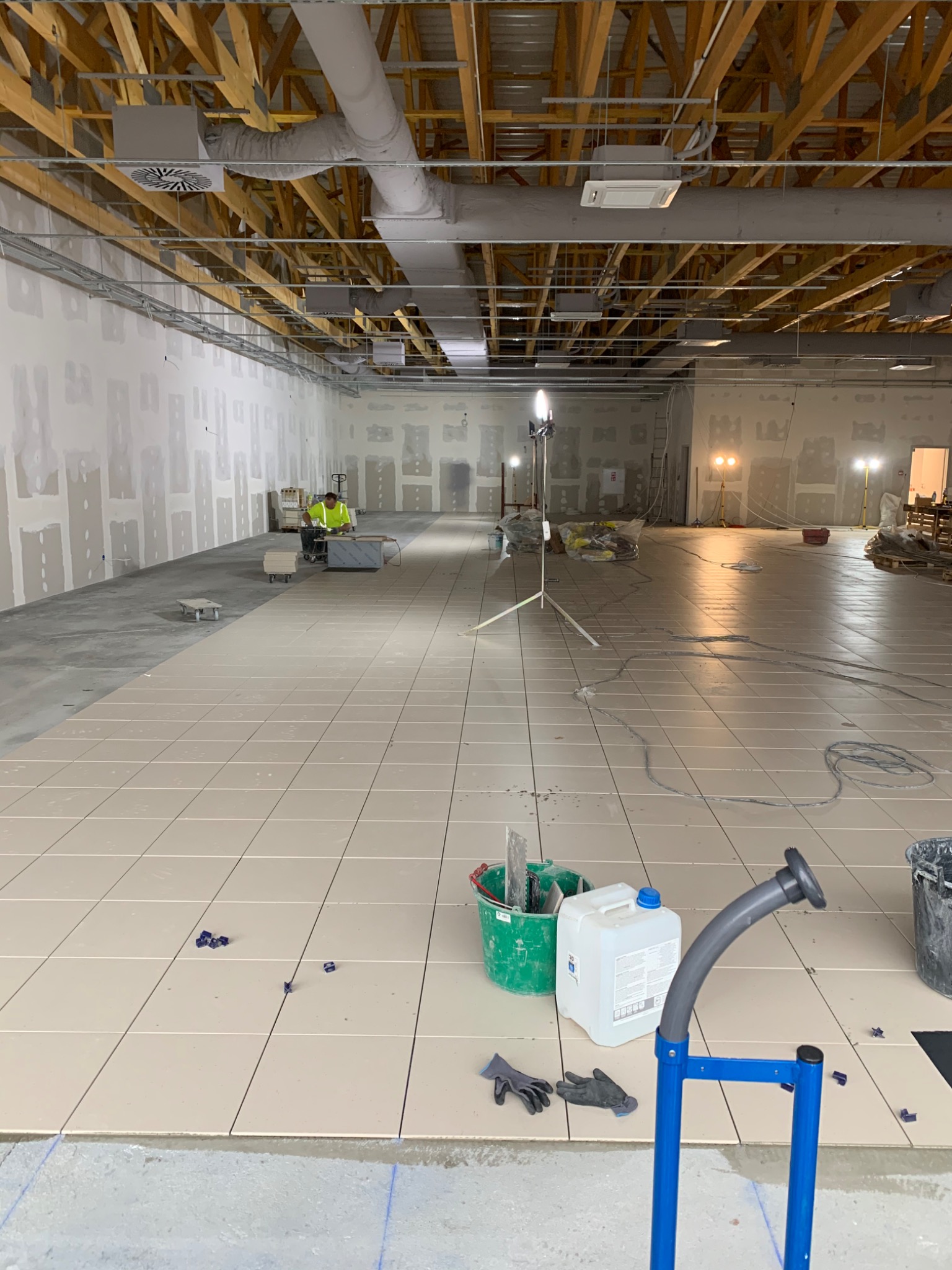 Układanie beżowych płytek podłogowych w dużym, surowym pomieszczeniu; widoczny pracownik w odblaskowej kamizelce, narzędzia glazurnicze i oświetlenie budowlane.