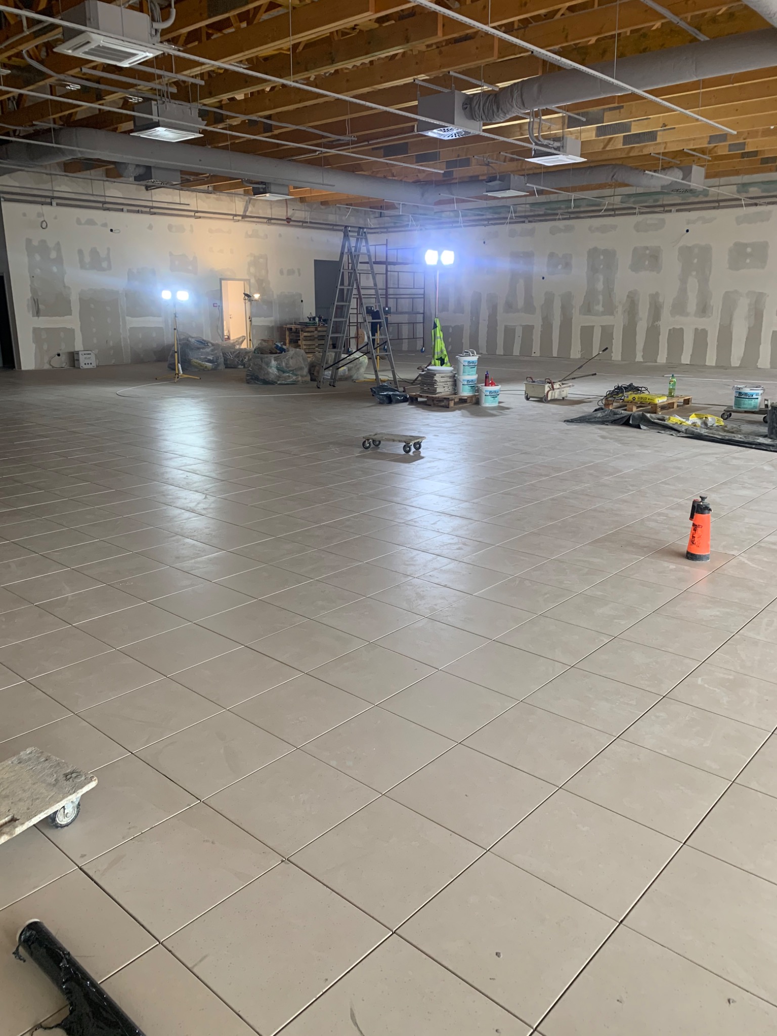 Rozległa przestrzeń w trakcie układania płytek podłogowych, widoczne narzędzia, materiały budowlane i drabina, ściany w trakcie przygotowania do malowania.