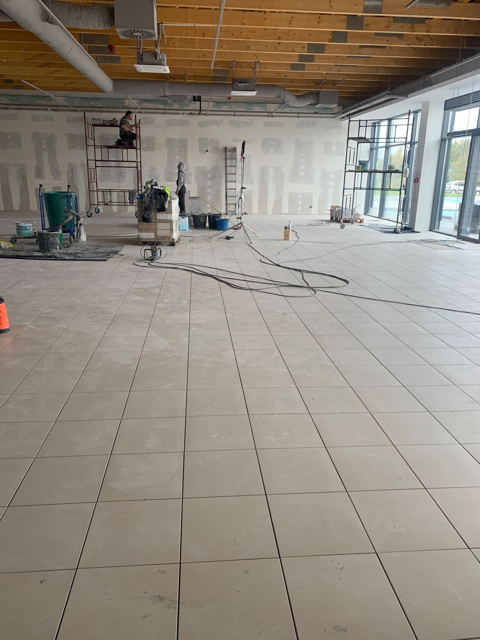 Układanie płytek podłogowych w dużym, jasnym pomieszczeniu; widoczni dwaj pracownicy, rusztowanie, narzędzia i materiały budowlane.