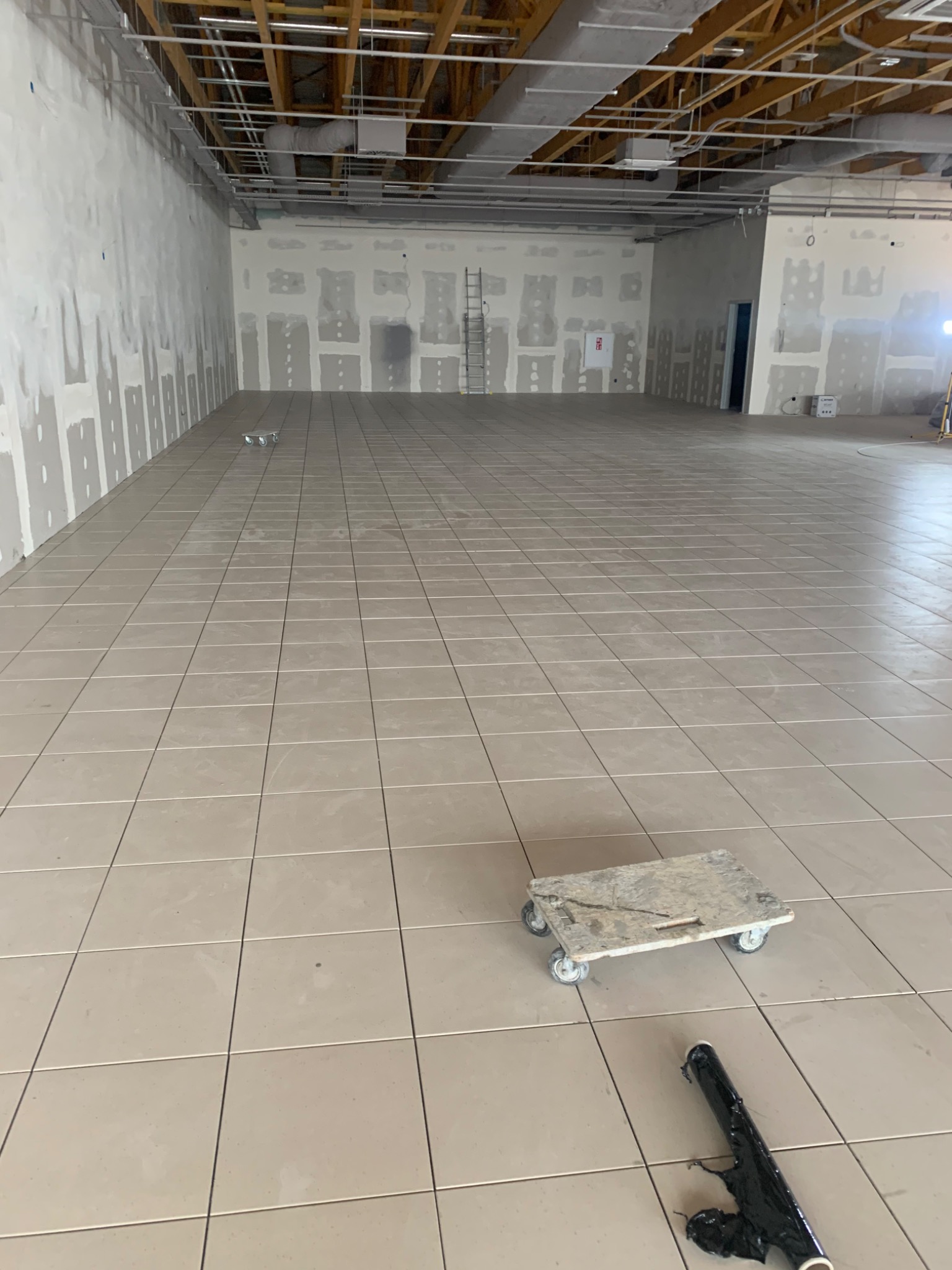 Beżowe płytki podłogowe ułożone w dużym, pustym pomieszczeniu w trakcie remontu, widoczny wózek transportowy i resztki folii ochronnej.