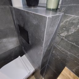ADMAR ADRIAN MARCISZ - Nowoczesna łazienka z szarą marmurową zabudową WC, drewnianą podłogą i czarnymi akcentami. Minimalistyczny design z dbałością o detale i wysoką jakość wykończenia.