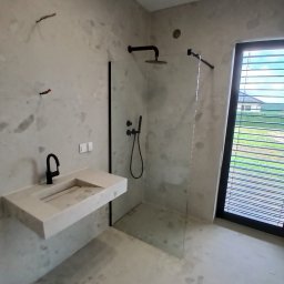 Admar Adrian Marcisz - Nowoczesna łazienka z podwieszaną umywalką z mikrocementu, czarną armaturą i kabiną walk-in. Ściany i podłoga w surowym stylu, duże okno z żaluzjami.